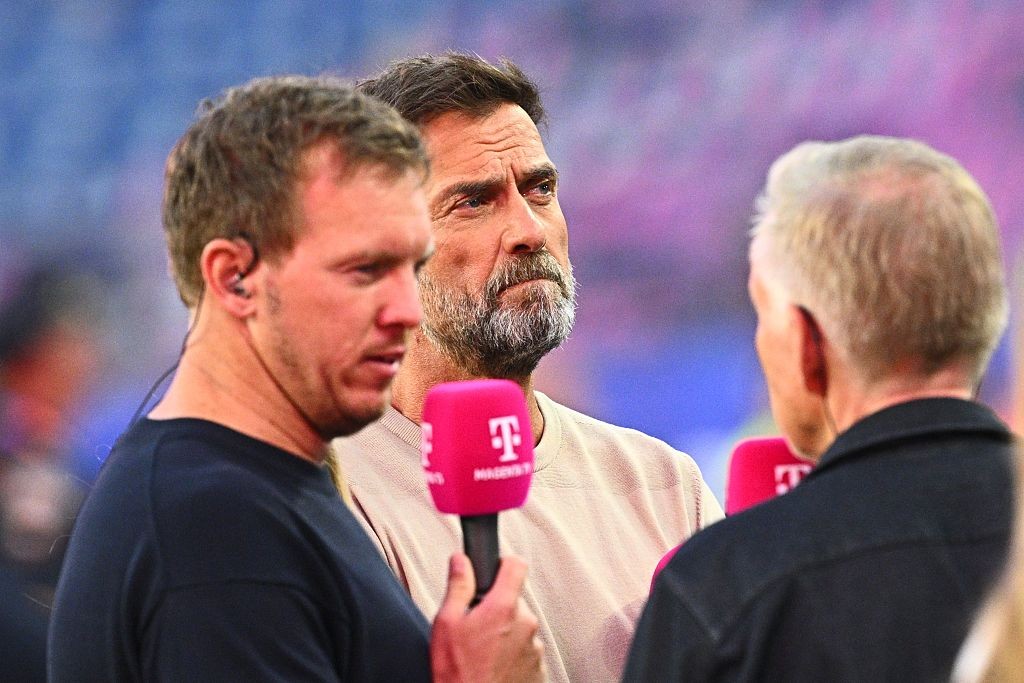 Três homens em um evento esportivo. O da esquerda, de camiseta preta e fone no ouvido, segura um microfone rosa com a letra T. O do meio, com barba grisalha, olha para a direita. O da direita está de costas, com cabelos grisalhos