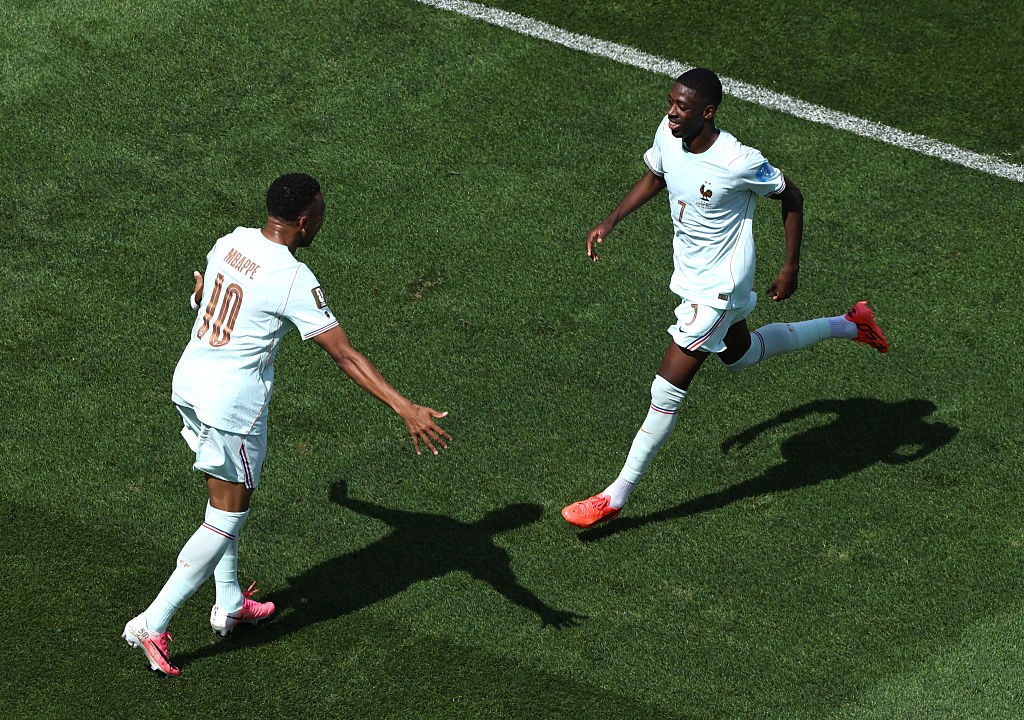 Dois jogadores de futebol com uniformes brancos e chuteiras coloridas correm em um campo de grama verde, um deles com o número 10 e o nome MBAPPE nas costas, estendendo o braço para o outro jogador que corre à frente com o número 7
