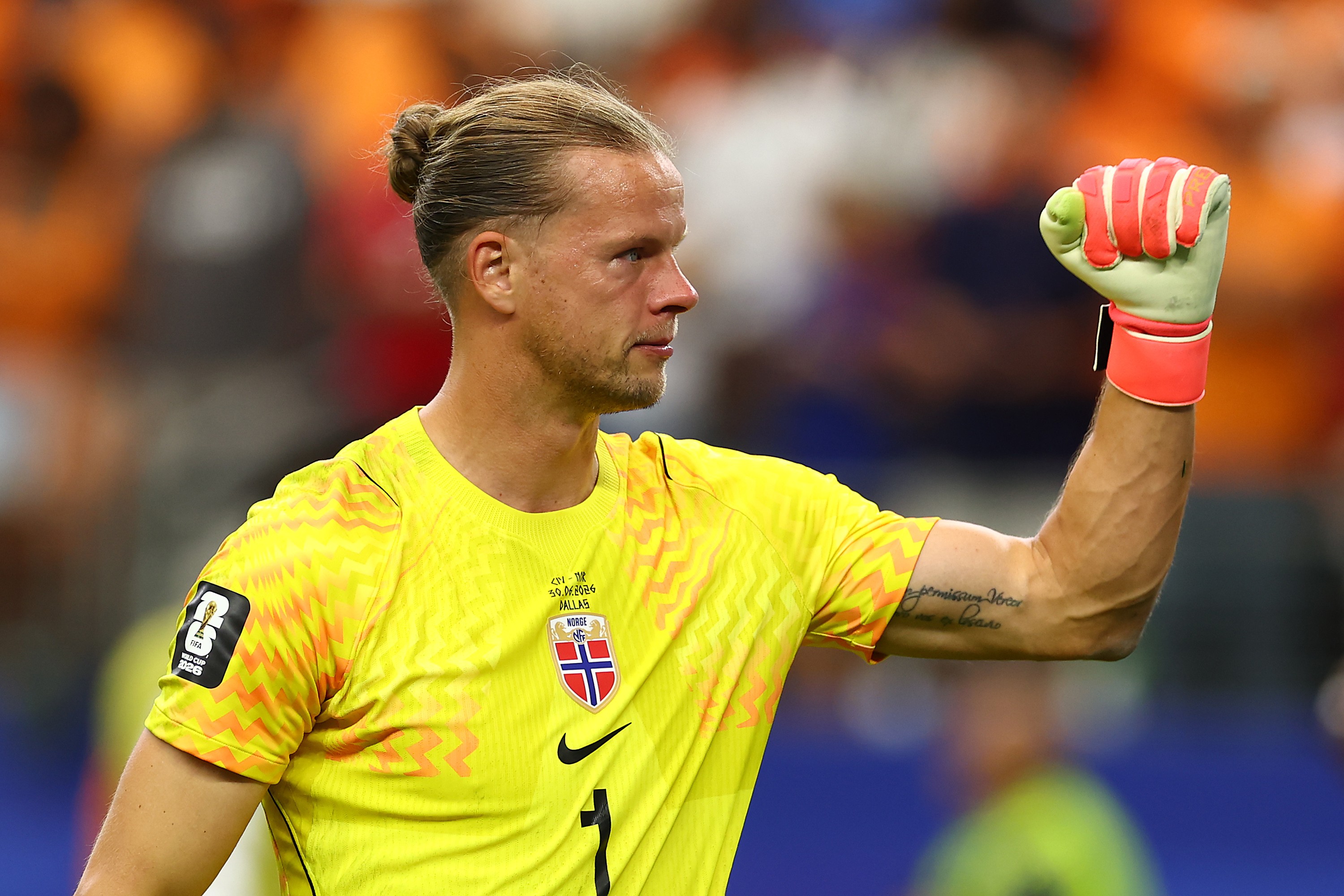 Goleiro de futebol com coque, camisa amarela e luvas verde-limão e laranja, com o punho esquerdo erguido em comemoração, em campo