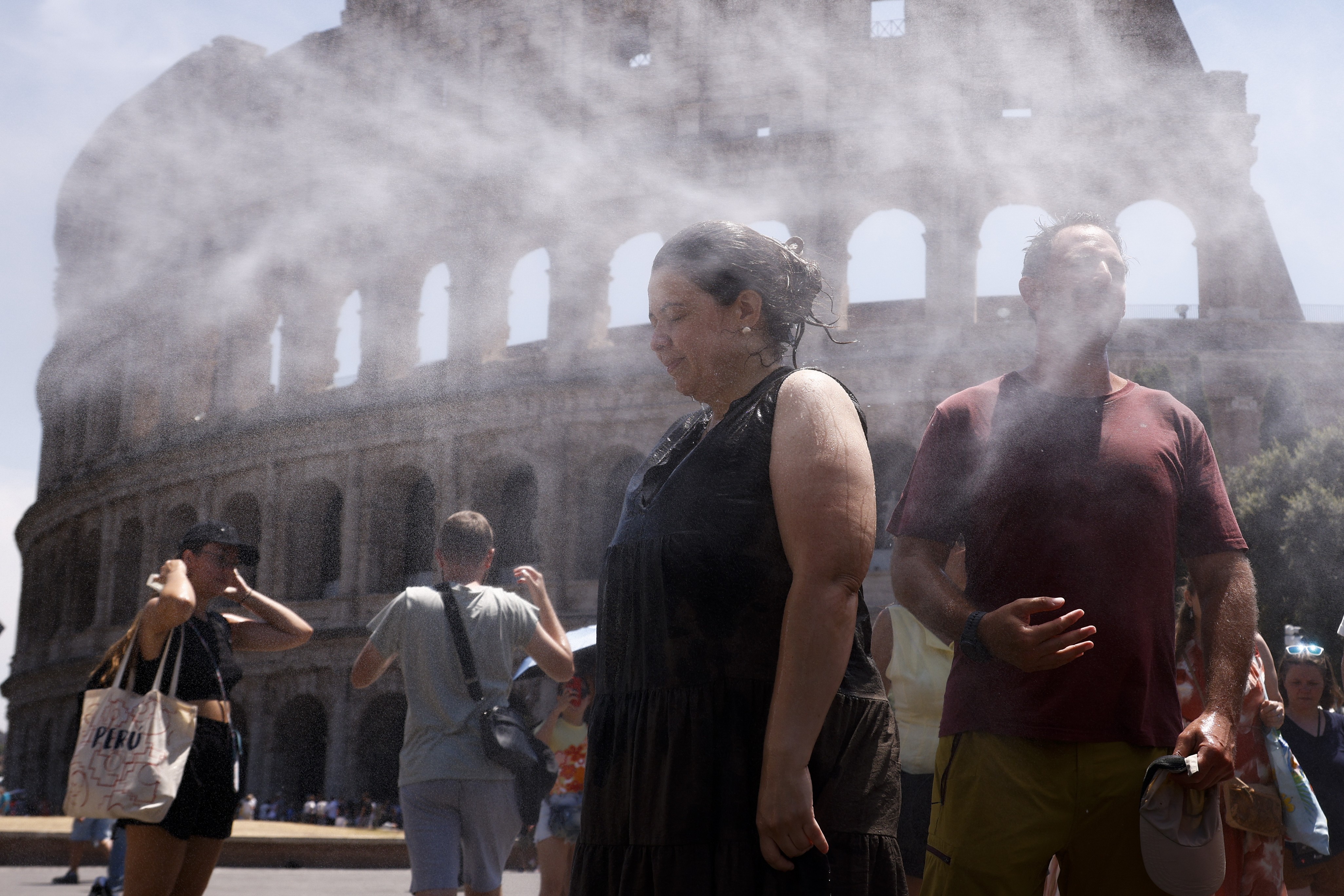 Pessoas recebem jatos de água para se refrescar em frente ao Coliseu, em Roma. Uma mulher de vestido preto e um homem de camiseta vinho estão no centro, com vapor de água ao redor. Outros turistas aparecem ao fundo, sob o sol forte e céu claro