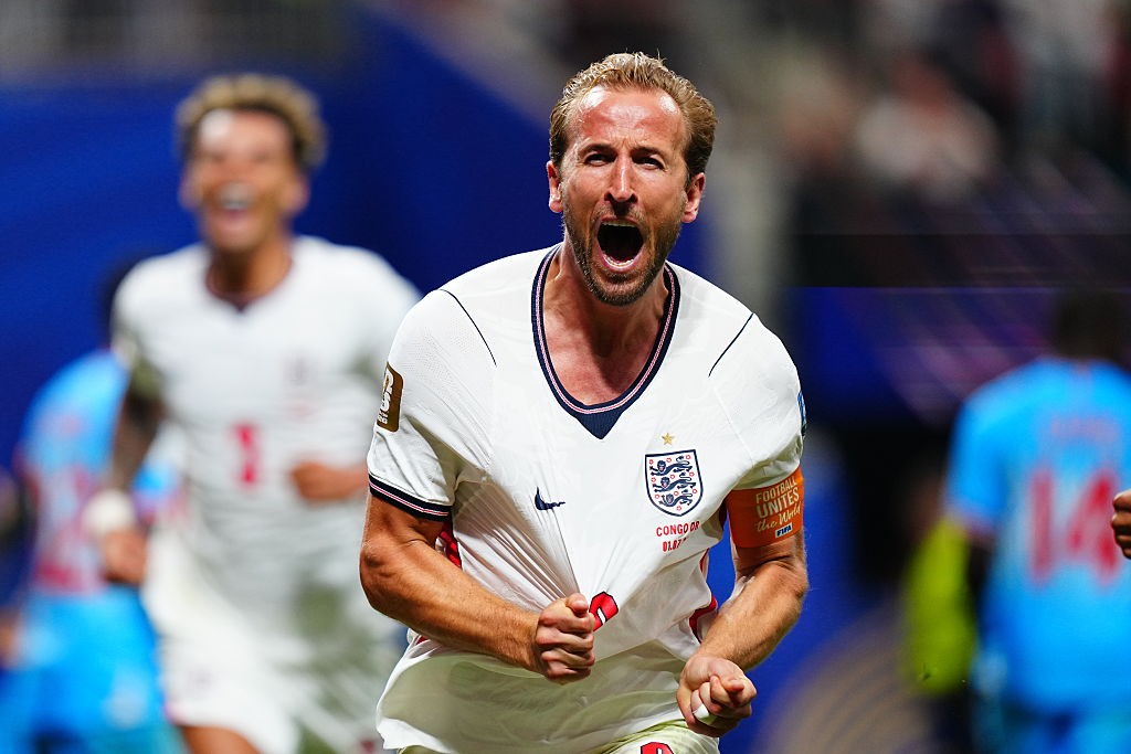Jogador de futebol Harry Kane, com camisa branca da Inglaterra, grita de alegria com a boca aberta, puxando a camisa. Outro jogador sorridente e desfocado aparece atrás, em campo de futebol