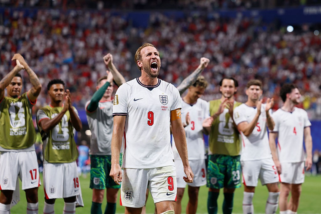 Harry Kane, camisa 9 da Inglaterra, grita e celebra com os braços abertos, enquanto outros jogadores aplaudem ao fundo em um estádio lotado