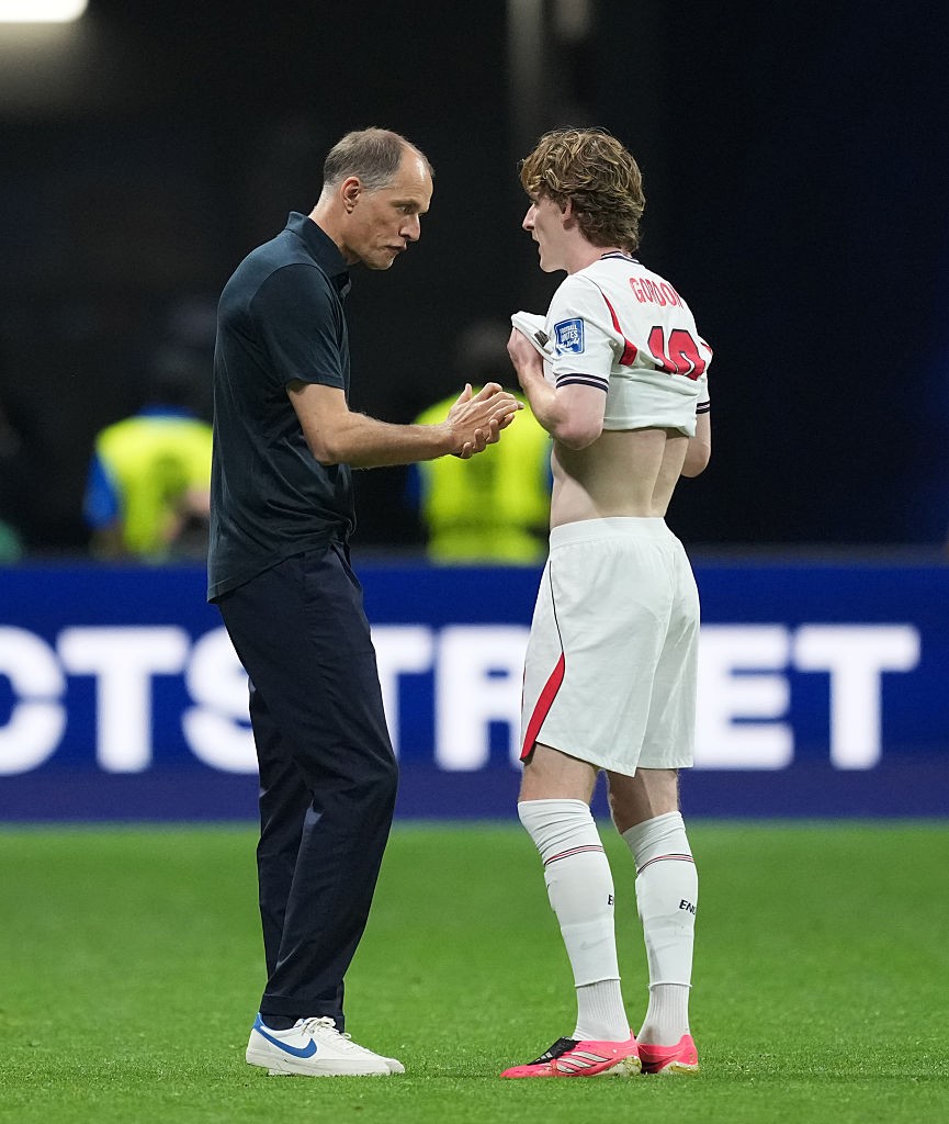 Dois homens em campo de futebol. Um técnico de camisa polo escura e calça preta conversa com um jogador de cabelo cacheado, que veste shorts brancos e segura a camisa branca com o nome GORDON e número 19.