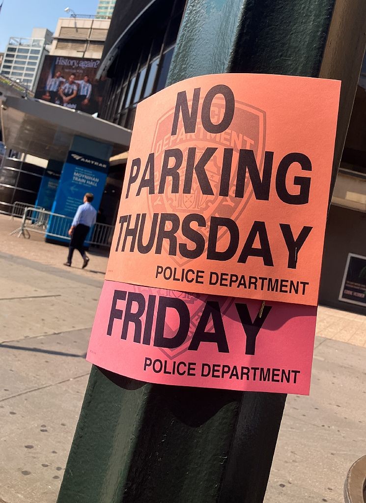 Dois avisos de NO PARKING (proibido estacionar) em laranja e rosa, fixados em um poste verde, com os dias THURSDAY (quinta-feira) e FRIDAY (sexta-feira) e a inscrição POLICE DEPARTMENT (Departamento de Polícia). Ao fundo, um homem caminha em frente à estação Moynihan Train Hall, com um outdoor exibindo a frase History again? (História de novo?)