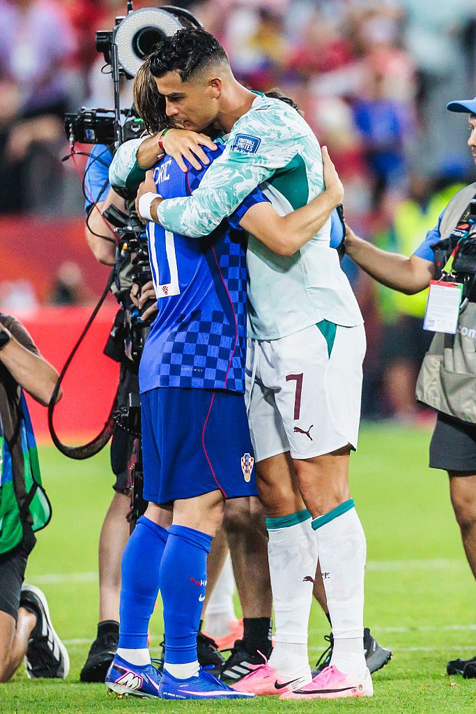 Cristiano Ronaldo, com camisa de manga longa azul-clara e branca, abra&ccedil;a um jogador de camisa azul xadrez e shorts azuis em campo de futebol, com c&acirc;meras e outros profissionais ao fundo