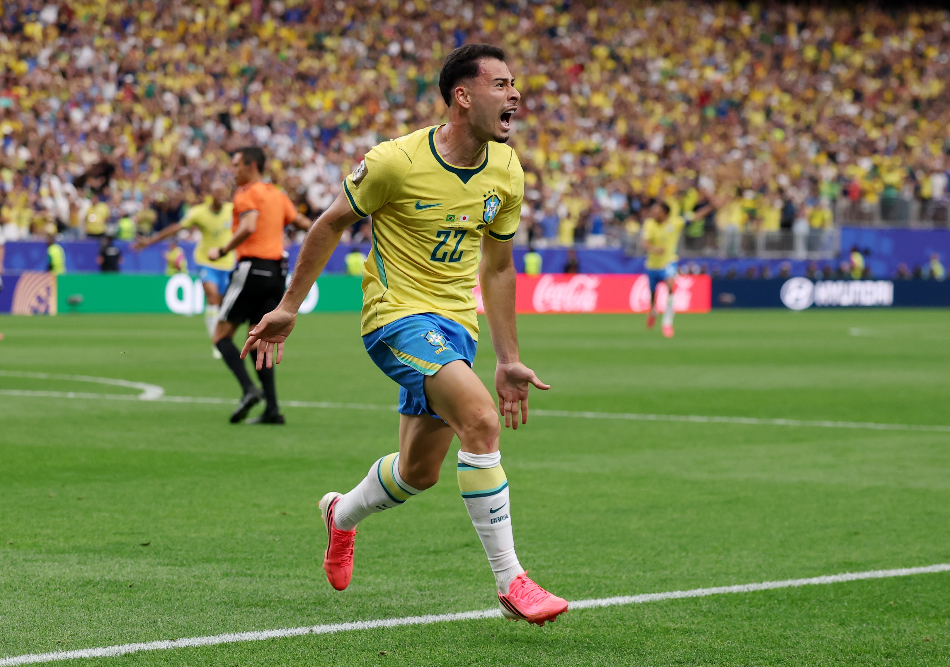 Jogador de futebol brasileiro com camisa amarela e shorts azuis, n&uacute;mero 22, corre no campo verde com a boca aberta em comemora&ccedil;&atilde;o, bra&ccedil;os abertos. Torcida amarela e azul ao fundo