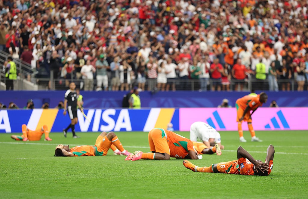 Jogadores de futebol de uniforme laranja e verde deitados no gramado, alguns com as mãos no rosto, em desespero após uma derrota. Um jogador de branco e outro de laranja estão curvados, e um de preto caminha ao fundo. A torcida lota as arquibancadas