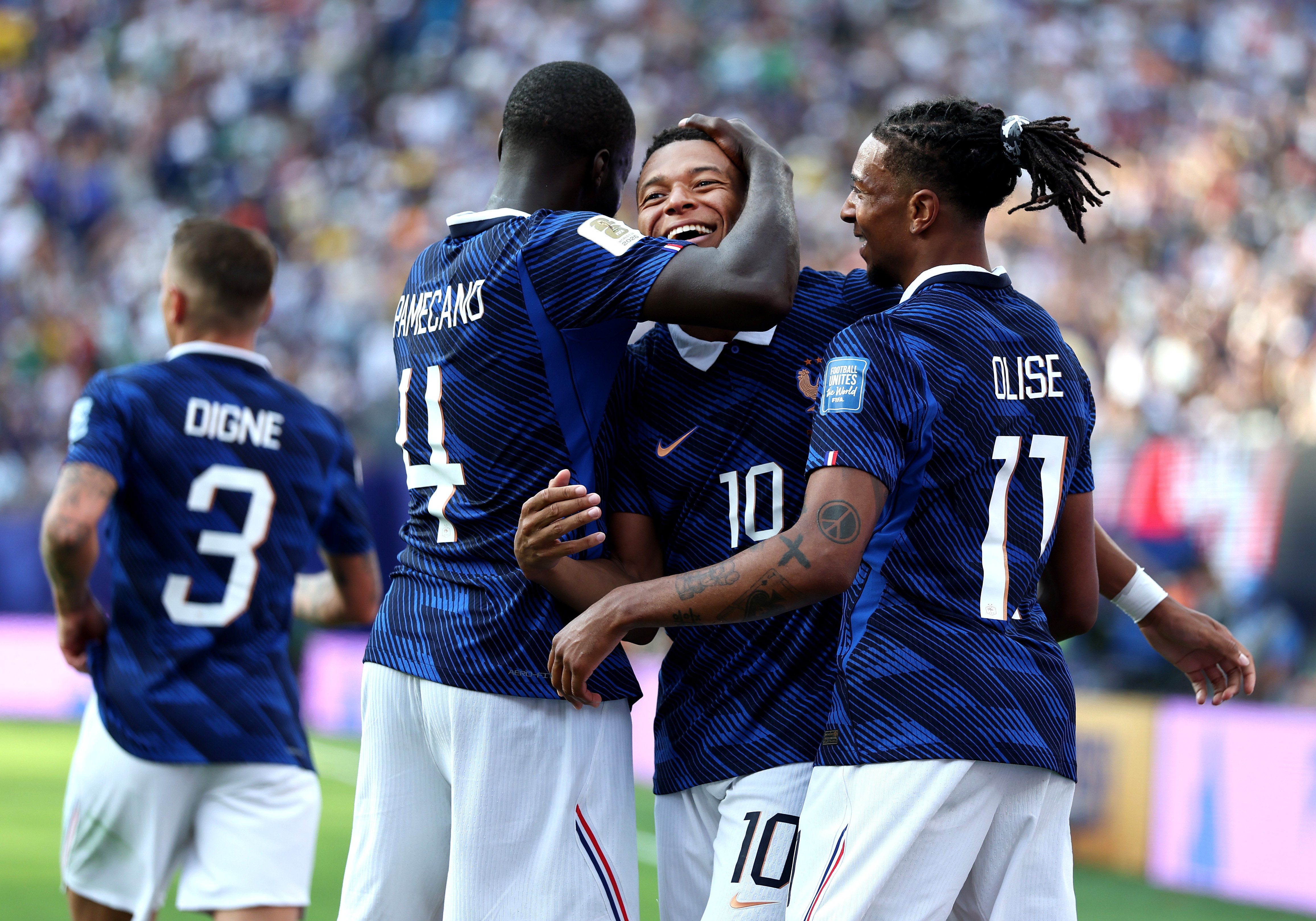 Jogadores de futebol franceses comemoram em campo. Um jogador de camisa 10 sorri, abraçado por outro de camisa 4. Um terceiro jogador de camisa 17, com dreadlocks presos, sorri ao lado. Todos vestem camisas azuis listradas e shorts brancos, em um estádio lotado