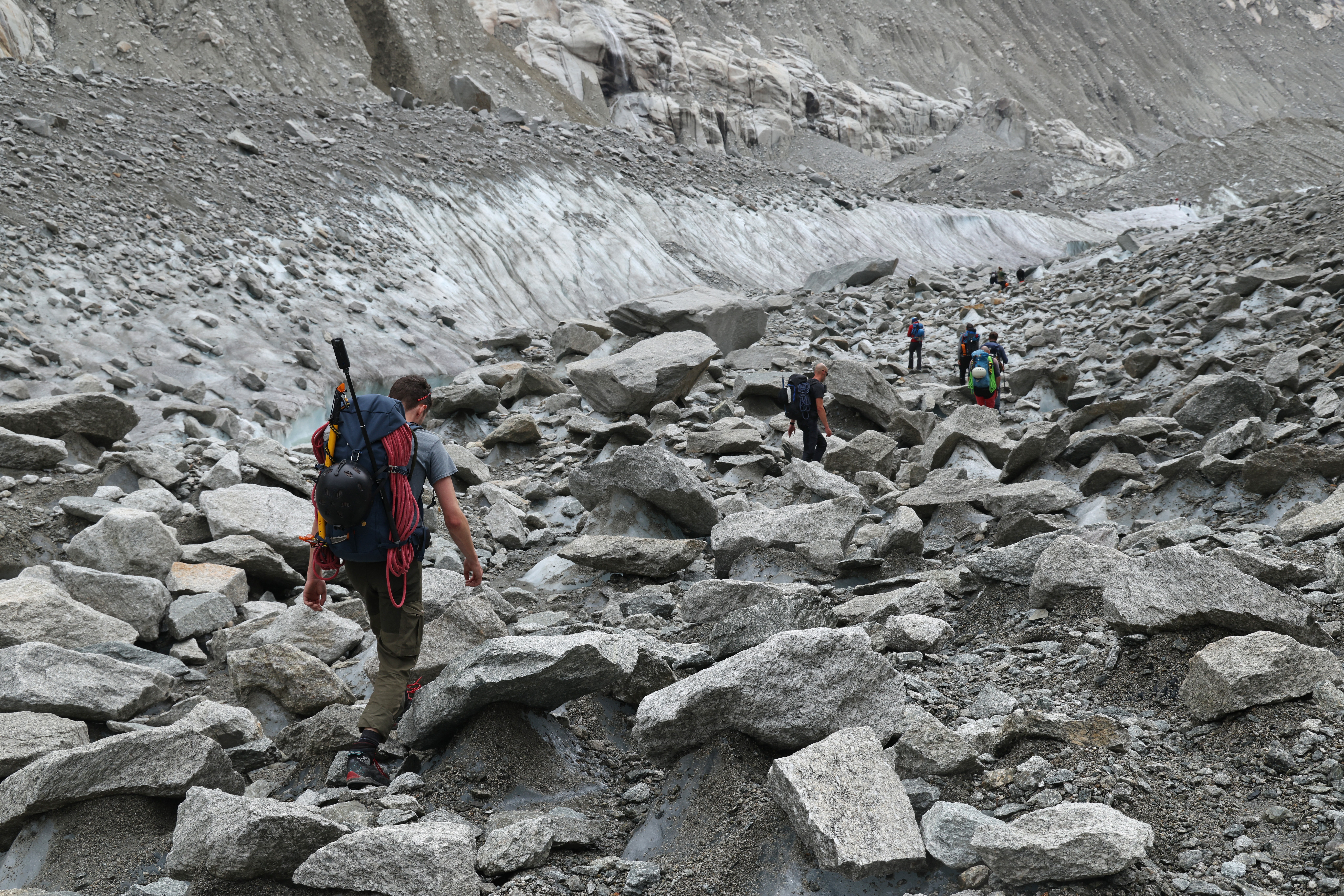 Um grupo de montanhistas caminha por um terreno rochoso e irregular, com um glaciar ao fundo. O homem em primeiro plano usa mochila grande e roupas de trilha, enquanto outros montanhistas seguem adiante. O cenário é árido, com muitas pedras e rochas de diferentes tamanhos, e o glaciar mostra camadas de gelo e detritos. O céu está nublado, e a atmosfera é de aventura e desafio.