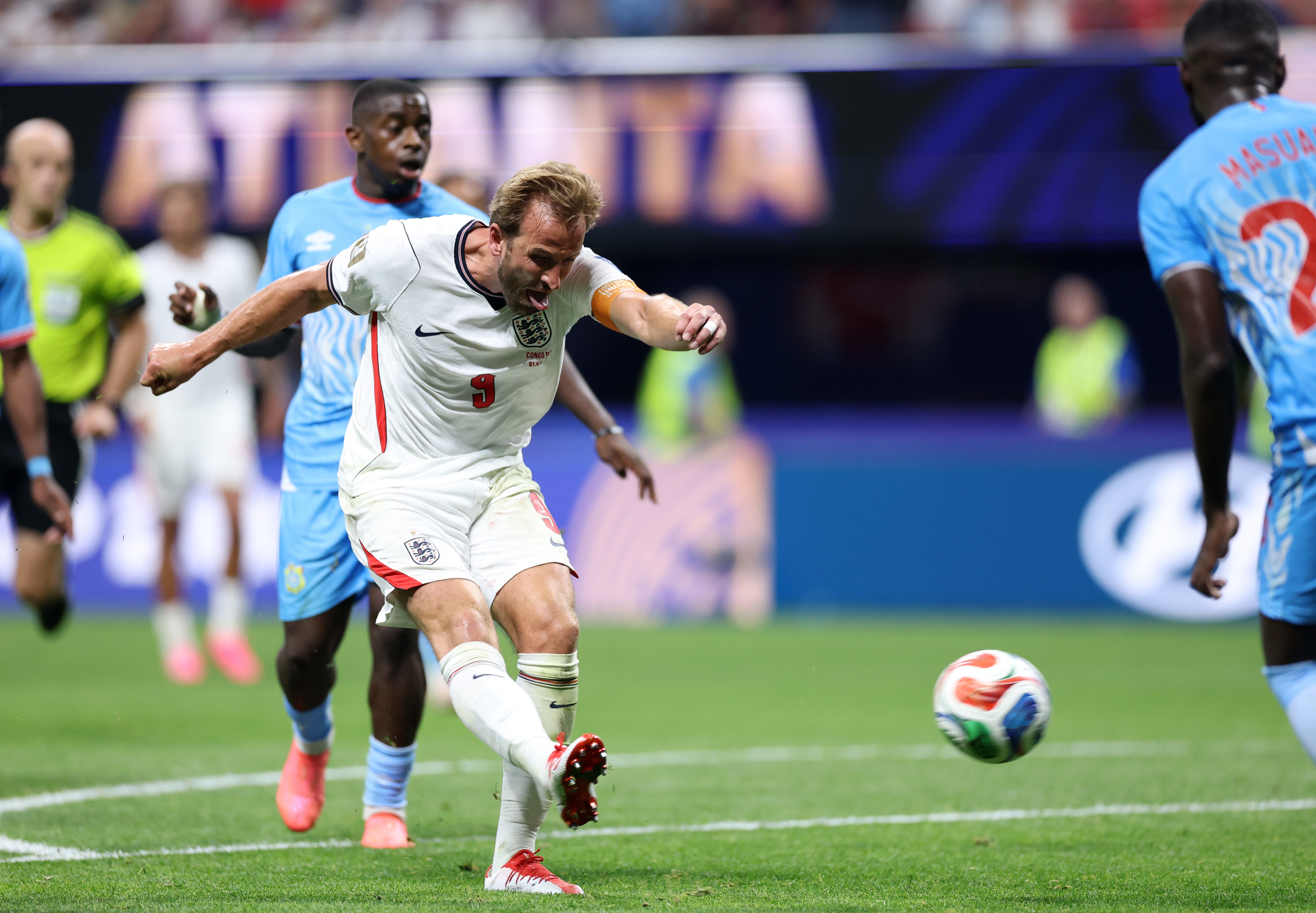 Jogador de futebol Harry Kane, com camisa branca da Inglaterra e número 9, chuta a bola com força em campo verde. Um jogador adversário de camisa azul observa atrás, em um estádio com luzes e público desfocado