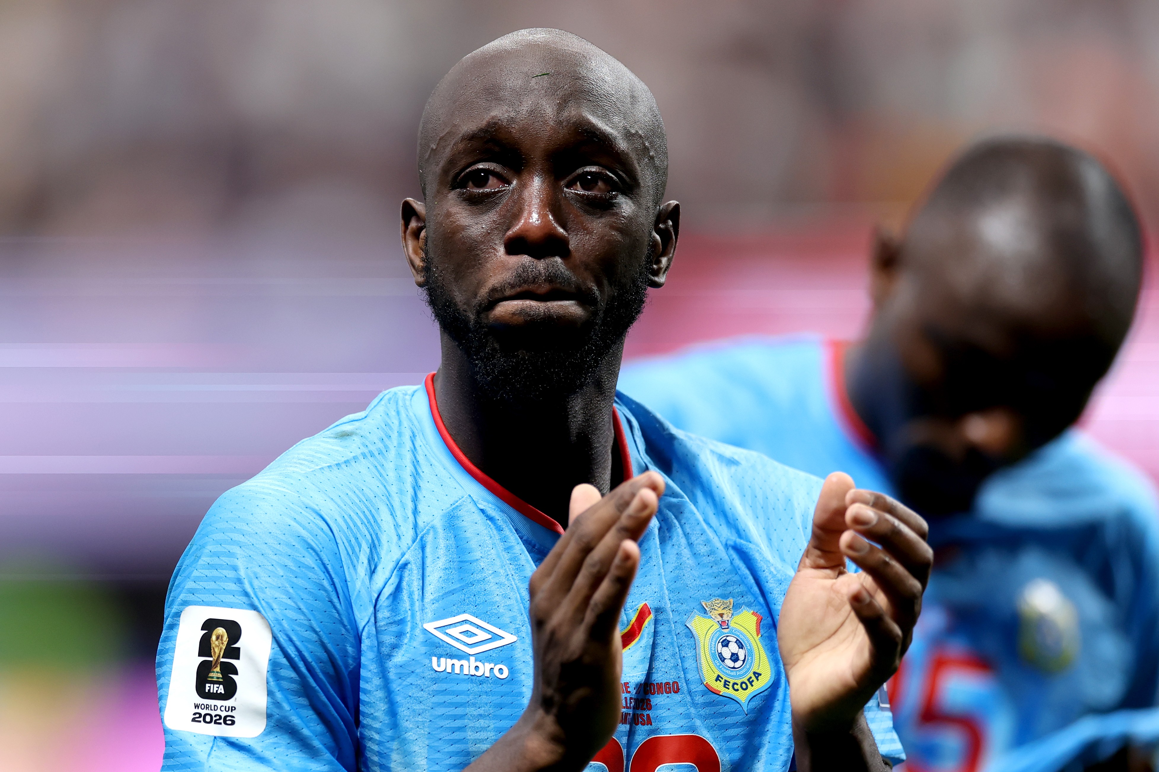 Homem negro careca, com barba rala, vestindo camisa de futebol azul-claro com detalhes vermelhos, aplaudindo com as mãos levantadas. No braço esquerdo, um patch da FIFA World Cup 2026. Outro jogador desfocado aparece ao fundo.
