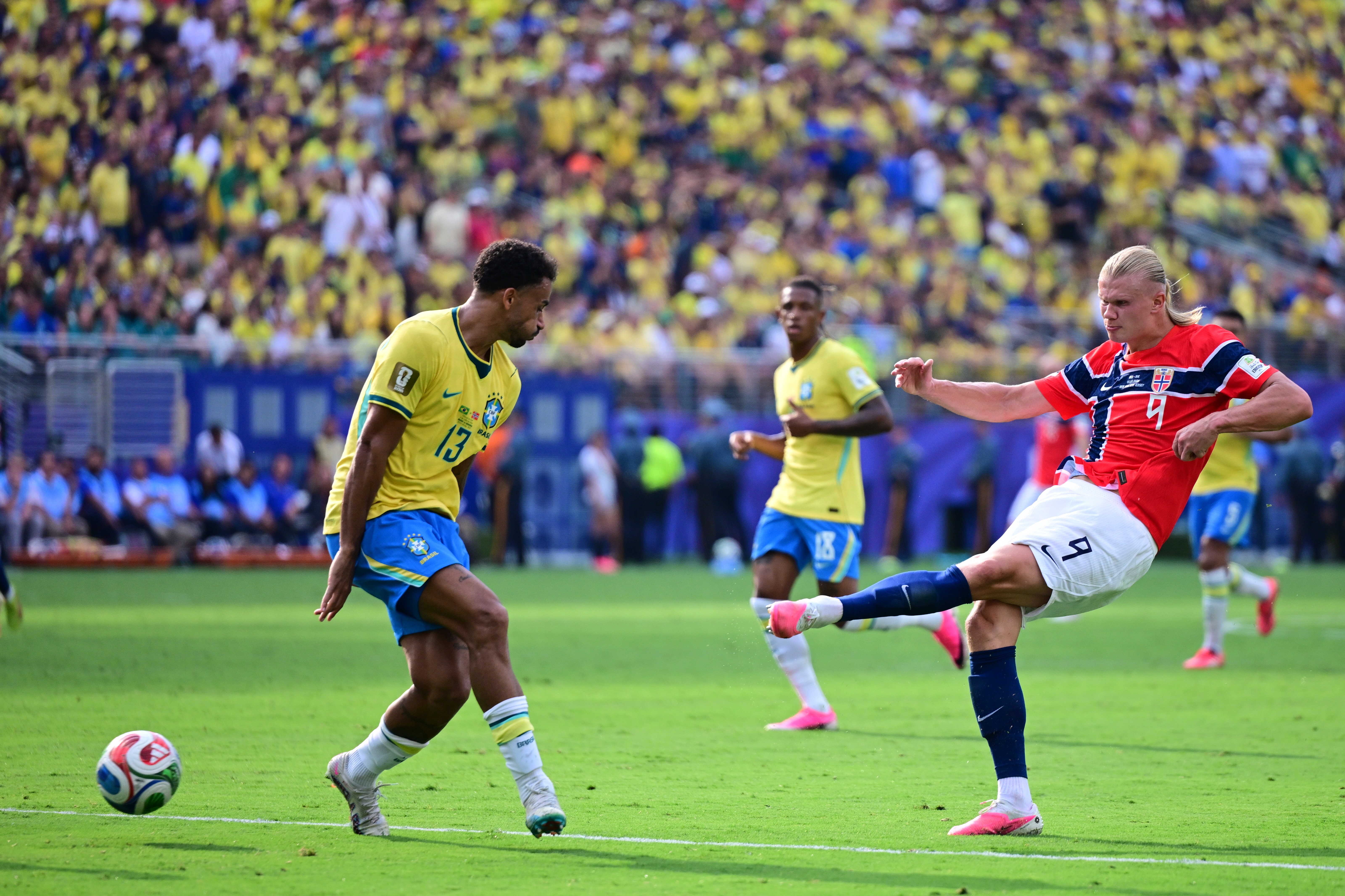 Dois jogadores de futebol em campo verde, um de camisa amarela do Brasil e outro de camisa vermelha da Noruega, disputam a bola. O jogador noruegu&ecirc;s chuta a bola com a perna direita, enquanto o brasileiro o observa. Torcedores em amarelo lotam as arquibancadas ao fundo