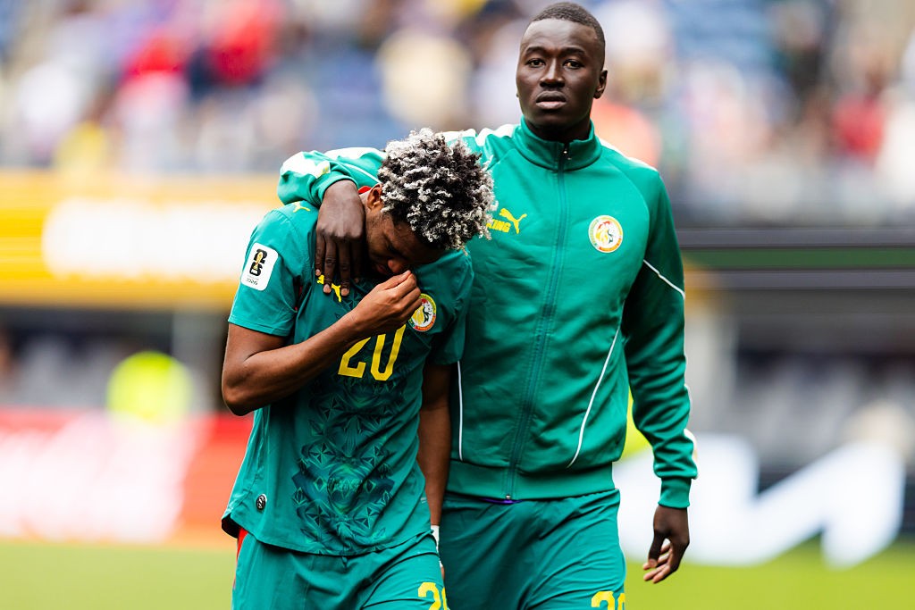 Dois jogadores de futebol negros em uniformes verdes. Um, com cabelo crespo claro, está cabisbaixo e com a mão no rosto, enquanto o outro o abraça pelo ombro, olhando para frente com expressão séria