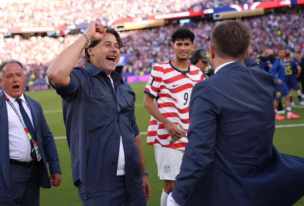 Três homens em um campo de futebol. Um homem de camisa azul escura e cabelo castanho escuro, com o punho cerrado e a boca aberta em comemoração. À esquerda, um homem de terno azul escuro e camisa branca, com a boca aberta. À direita, um jogador de camisa listrada vermelha e branca com o número 9, olhando para o homem de terno azul de costas. Ao fundo, uma multidão em um estádio