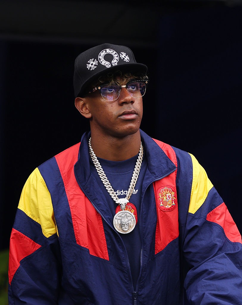 Homem jovem de pele escura, boné preto com símbolos brancos, óculos de armação dourada e lentes claras, corrente grossa prateada com pingente grande, jaqueta esportiva azul, vermelha e amarela, com emblema da Espanha no peito, olhando para o lado direito com expressão séria