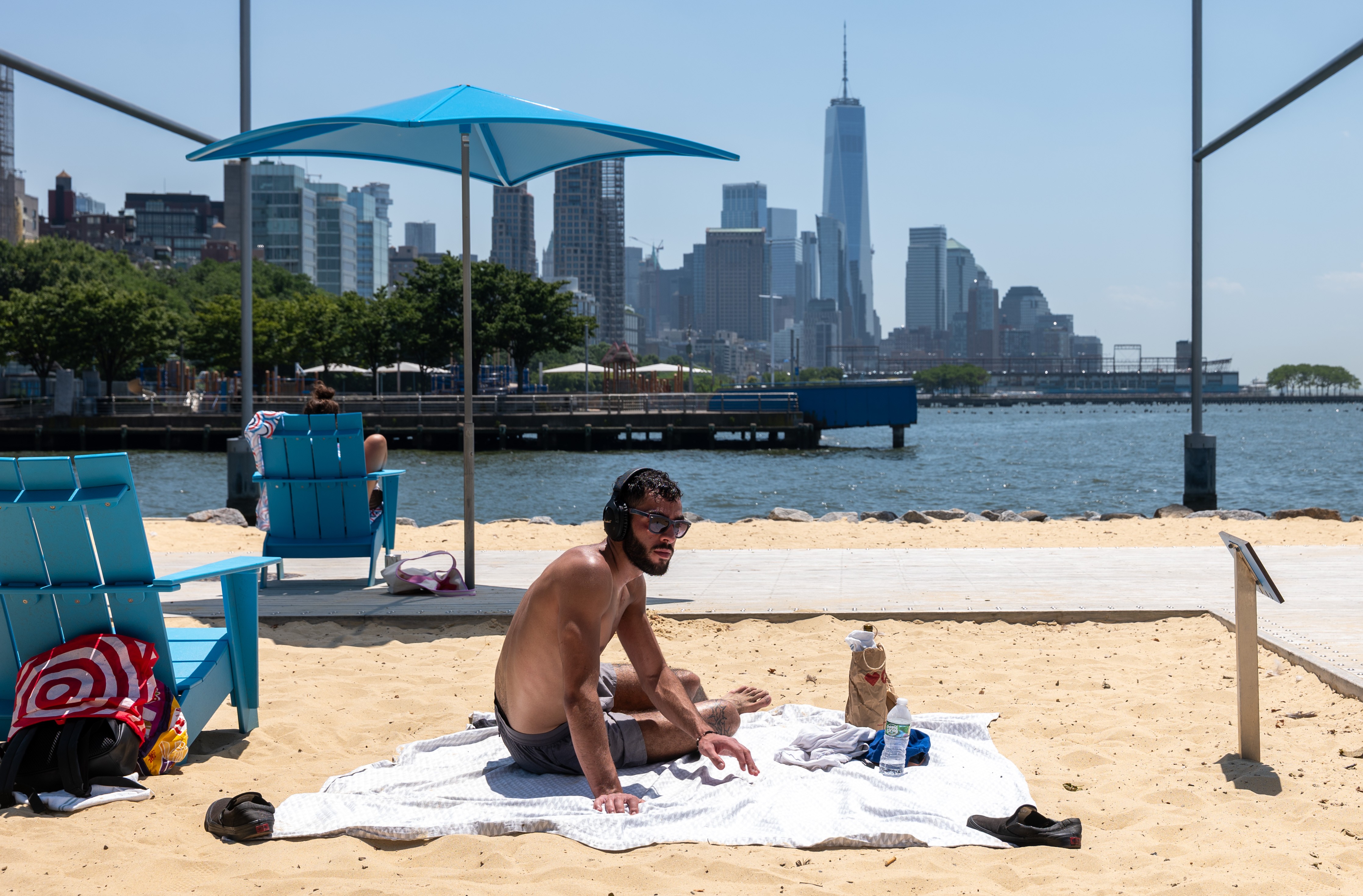 Homem de &oacute;culos escuros e fones de ouvido sentado em uma toalha branca na areia, com a silhueta de Nova York ao fundo, sob um c&eacute;u azul claro