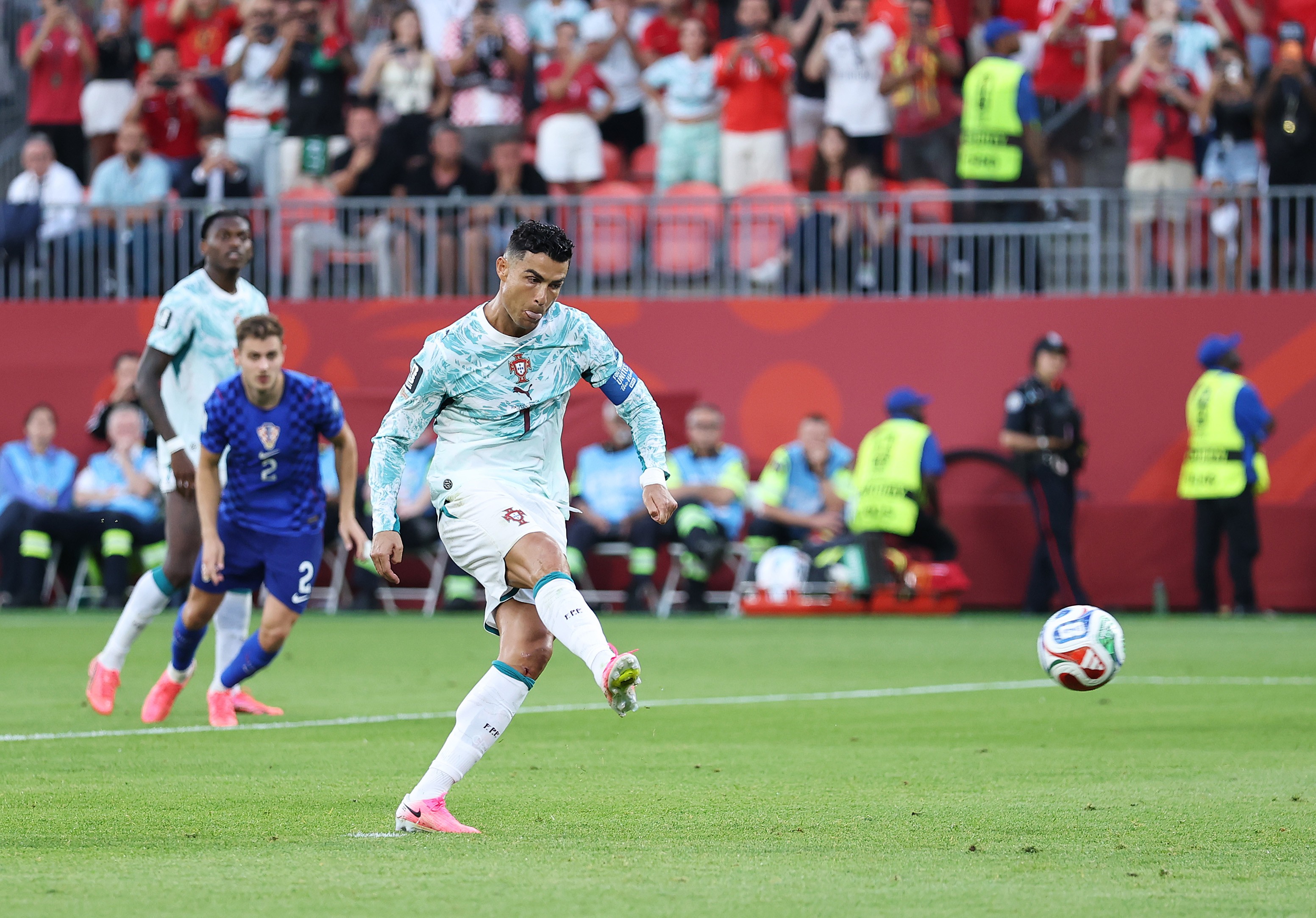 Cristiano Ronaldo, com camisa branca e azul-claro, chuta uma bola de futebol no campo verde. Outros jogadores e torcedores em segundo plano.