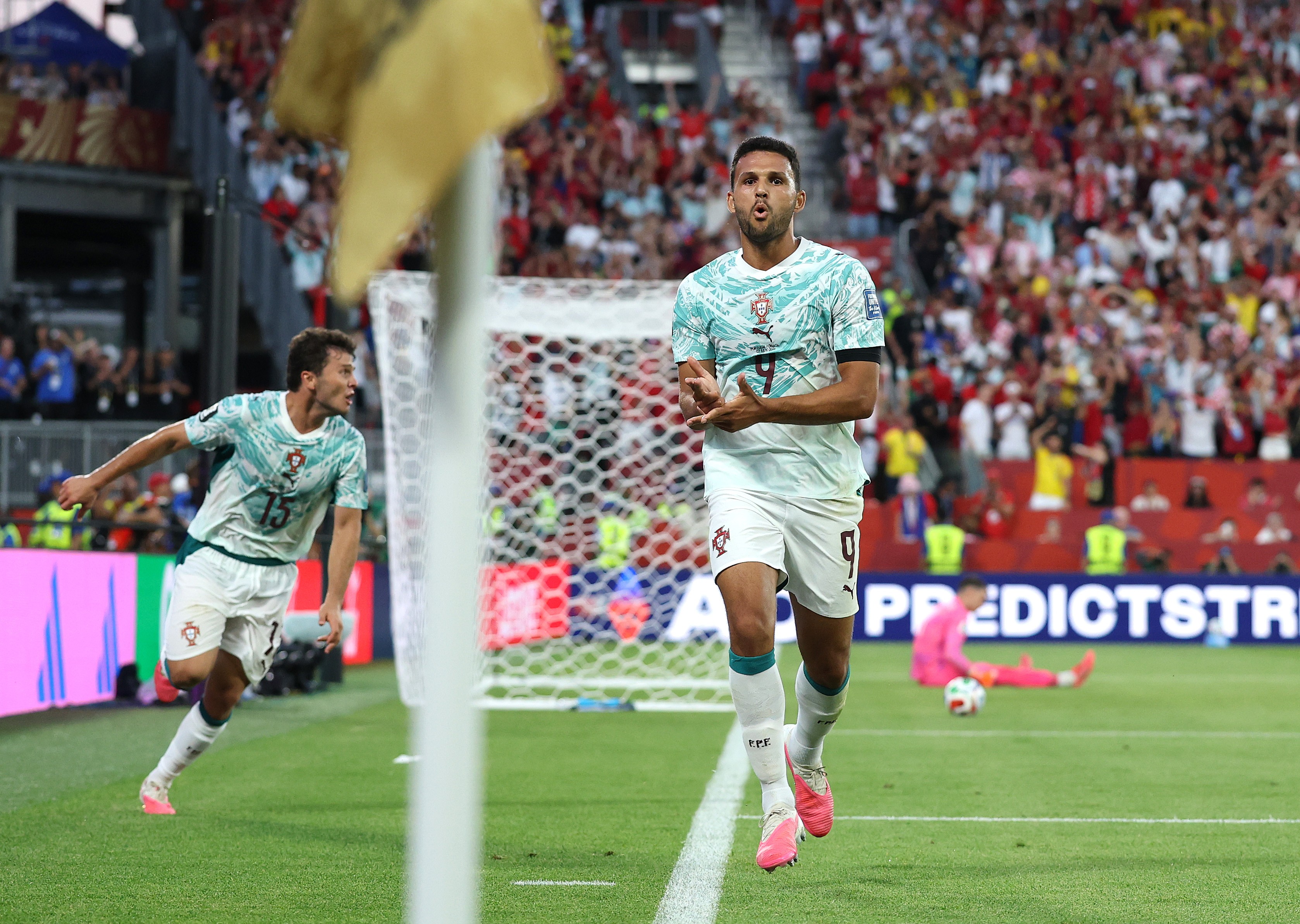 Dois jogadores de futebol portugueses, um com a camisa 9 e outro com a 15, comemoram um gol em campo. O jogador da camisa 9 est&aacute; mais pr&oacute;ximo, com as m&atilde;os unidas e boca aberta, enquanto o da 15 corre ao fundo. A torcida lota as arquibancadas e um goleiro de rosa est&aacute; ca&iacute;do no ch&atilde;o. Uma bandeira de escanteio desfocada aparece em primeiro plano