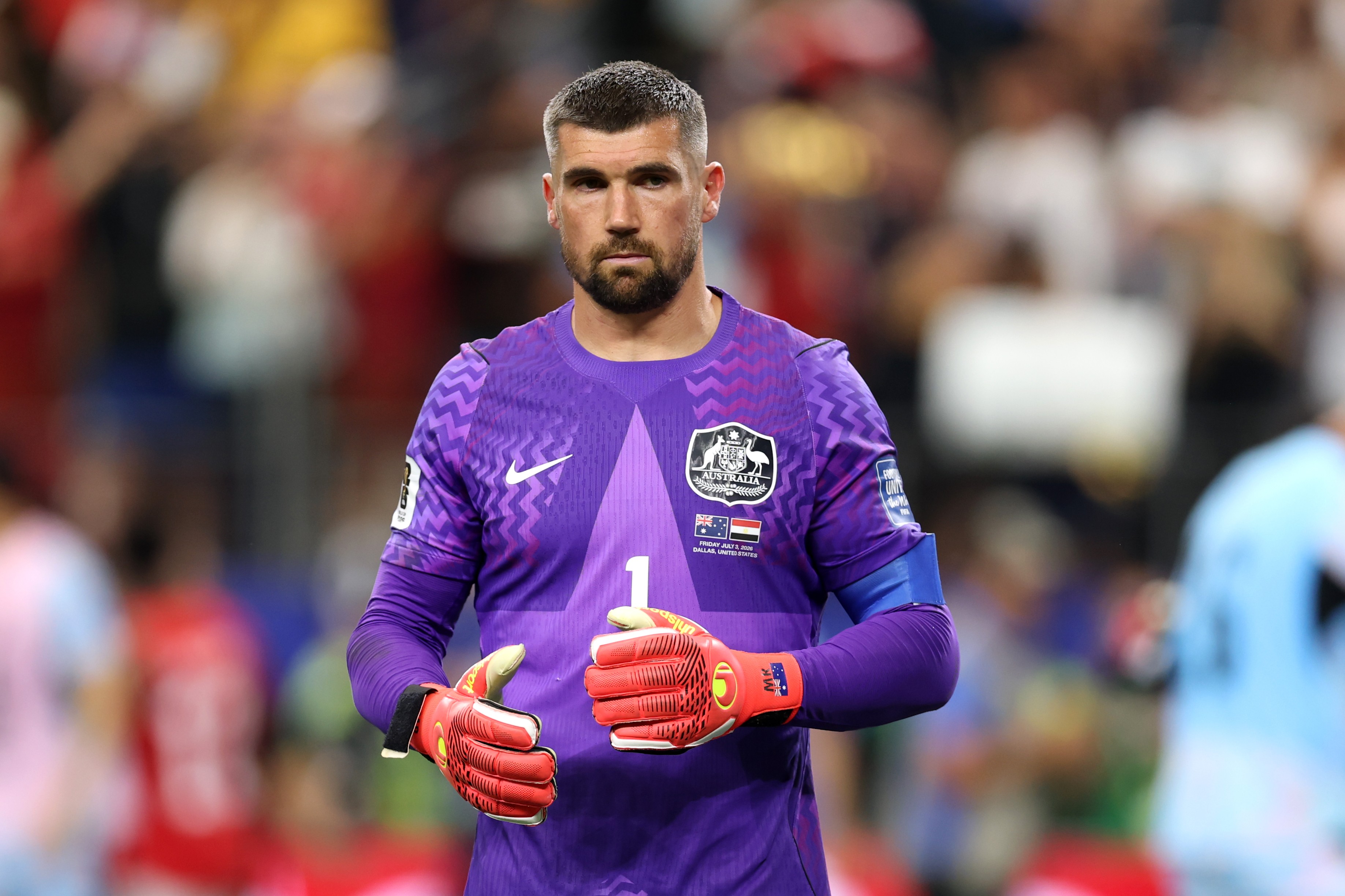 Goleiro com barba e cabelo curto, vestindo camisa roxa de manga longa com detalhes em zigue-zague e luvas de goleiro vermelhas, em campo de futebol