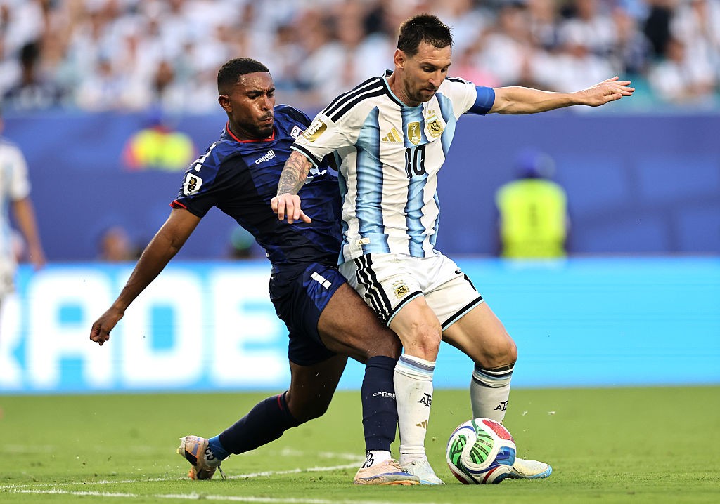 Dois jogadores de futebol em campo. Um jogador de camisa azul e calção azul tenta desarmar um jogador de camisa listrada azul e branca com o número 10, que está com a bola nos pés. Ambos estão em movimento, com o jogador de camisa azul se inclinando para frente e o de camisa listrada com o braço estendido para o lado, mantendo o equilíbrio. O gramado verde e a arquibancada desfocada ao fundo completam a cena