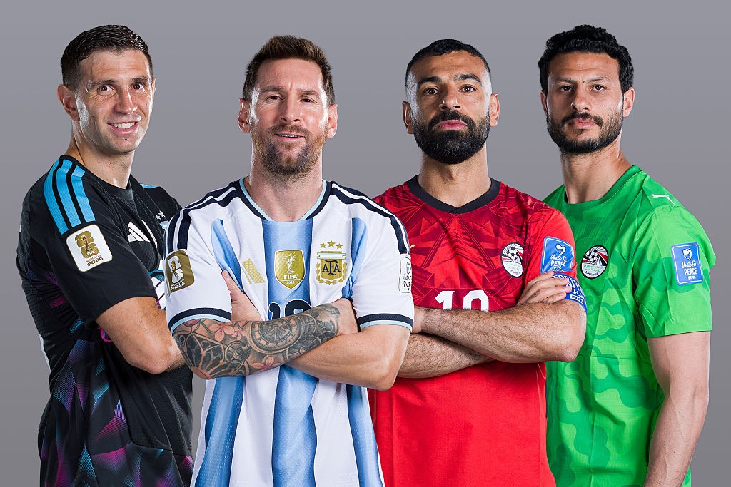 Quatro jogadores de futebol, da esquerda para a direita: Emiliano Martínez sorrindo em uniforme de goleiro preto e azul, Lionel Messi com camisa argentina listrada de branco e azul claro e braços cruzados, Mohamed Salah com camisa vermelha e braços cruzados, e um jogador egípcio com camisa verde
