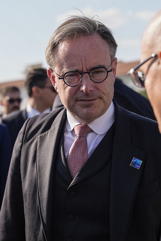 Homem de meia-idade, cabelos grisalhos e óculos redondos, vestindo terno escuro, colete e gravata rosa estampada, com um broche azul no paletó, conversando com outra pessoa em ambiente externo