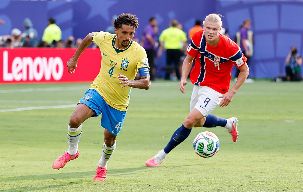 Dois jogadores de futebol correm no campo verde. Um, à esquerda, veste camisa amarela e shorts azuis do Brasil, com o número 4. O outro, à direita, usa camisa vermelha e branca da Noruega, com o número 9, e chuta uma bola colorida. Ao fundo, uma placa vermelha com o logo Lenovo em branco.