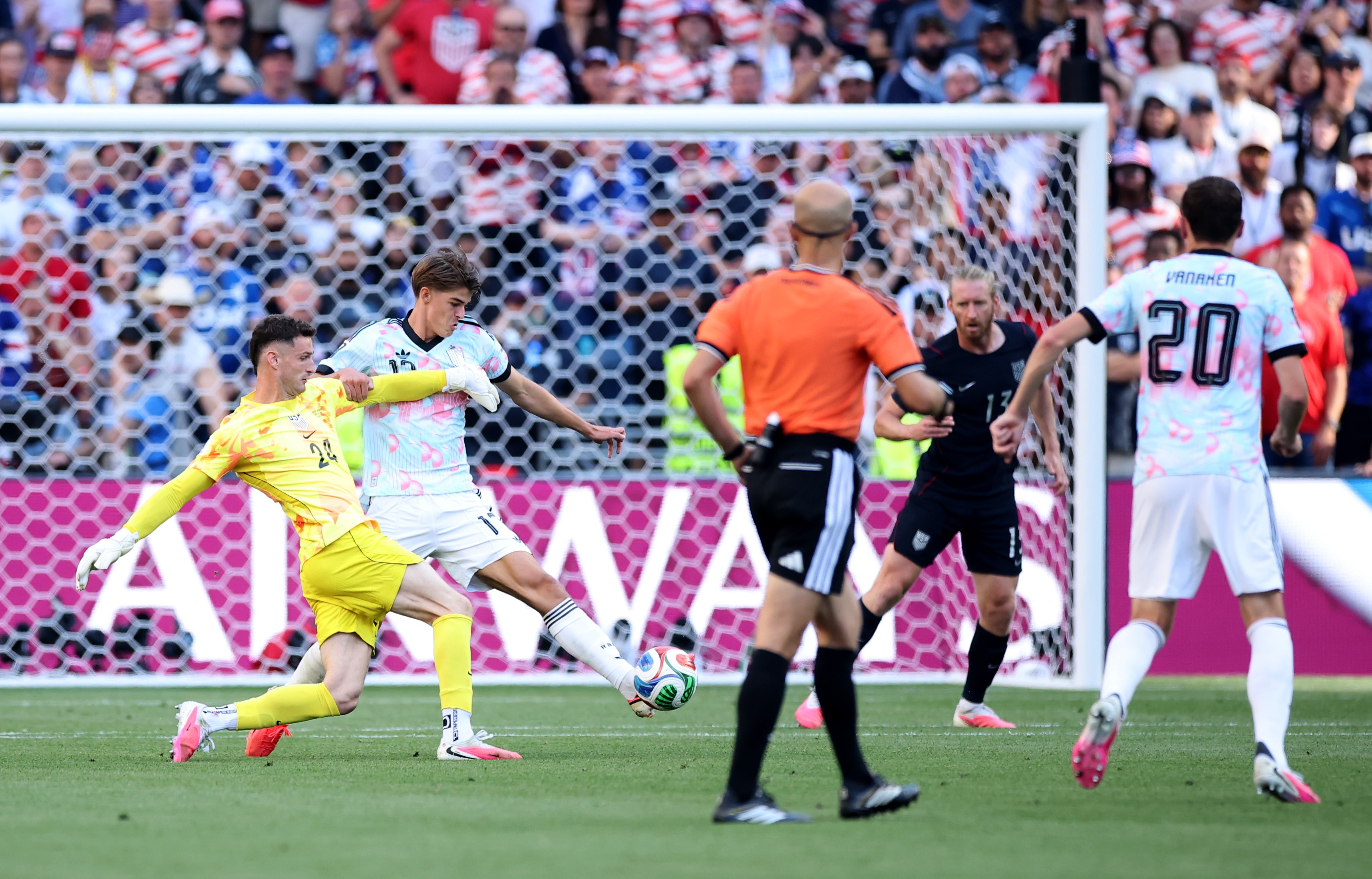 Goleiro de camisa amarela e jogador de camisa branca e rosa disputam a bola no campo de futebol, com o gol ao fundo. Um &aacute;rbitro de laranja e outros jogadores observam a jogada. A torcida lota as arquibancadas.