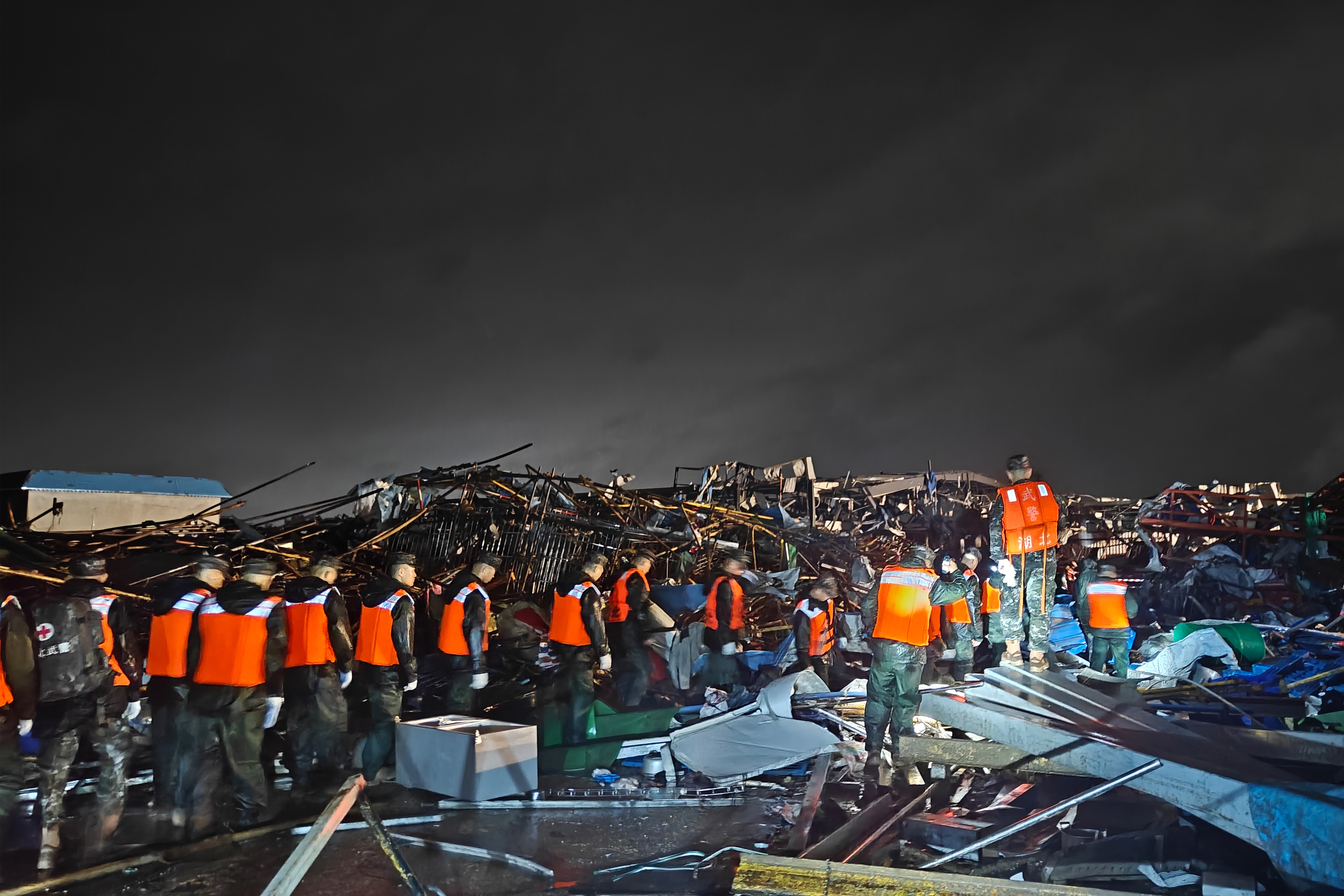 Equipe de resgate com coletes laranja e uniformes escuros caminha sobre escombros de uma &aacute;rea devastada, sob c&eacute;u noturno nublado