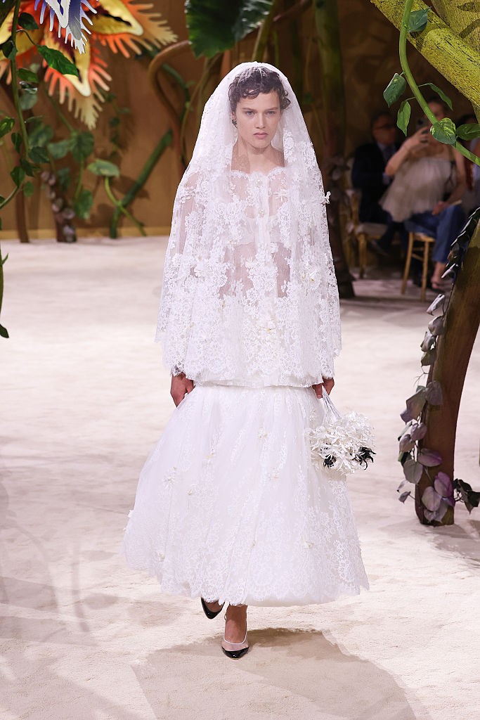 Modelo com cabelo cacheado escuro, usando um vestido de noiva branco rendado com véu e um buquê de flores brancas, desfila em passarela clara com decoração de folhagens e árvores ao fundo