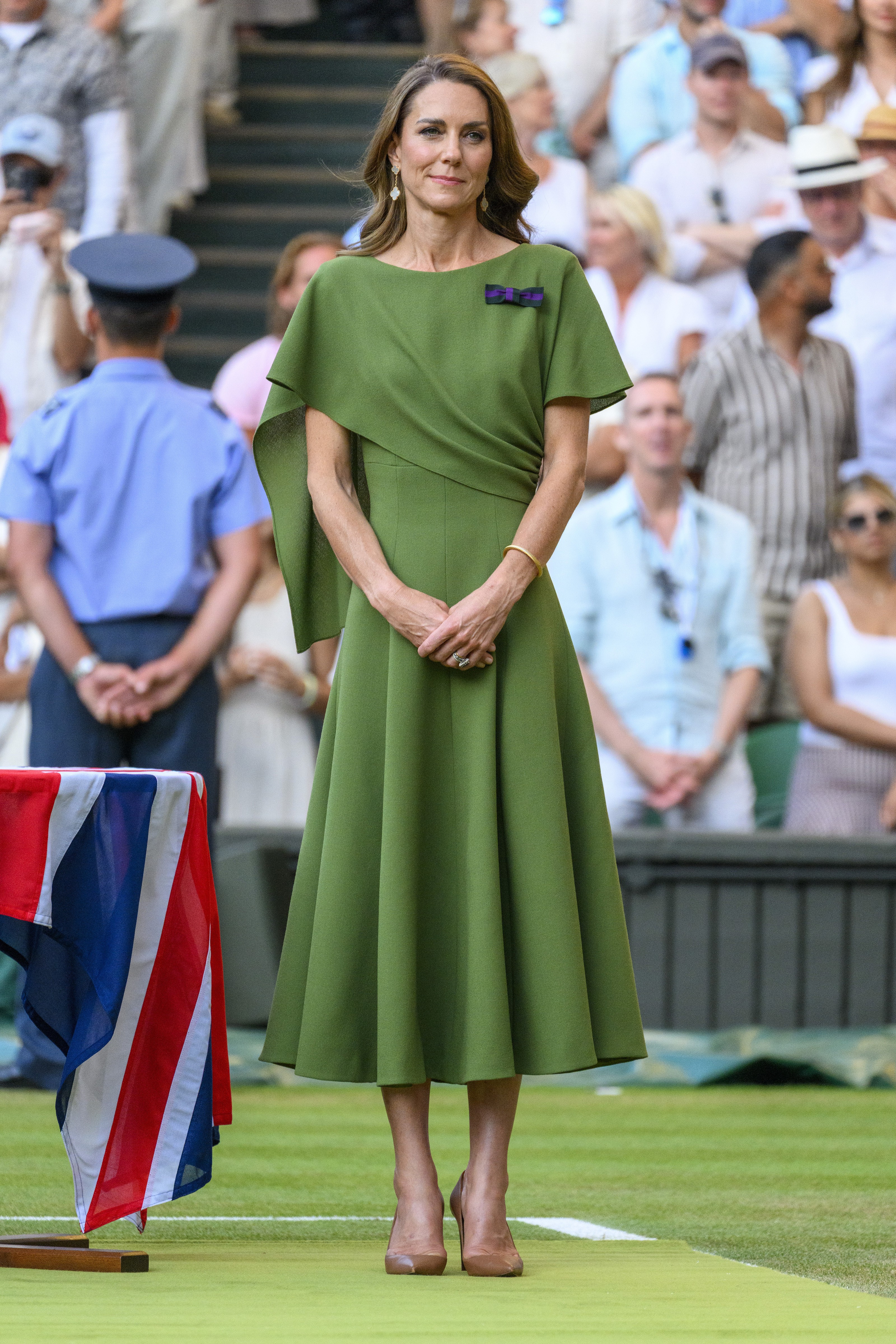 Mulher de cabelos castanhos, vestindo um elegante vestido verde-oliva com detalhes drapeados e um broche roxo, está de pé com as mãos cruzadas, em um evento ao ar livre com público ao fundo. Ela usa brincos e uma pulseira dourada, e sapatos de salto nude. À esquerda, uma bandeira do Reino Unido está sobre uma mesa.