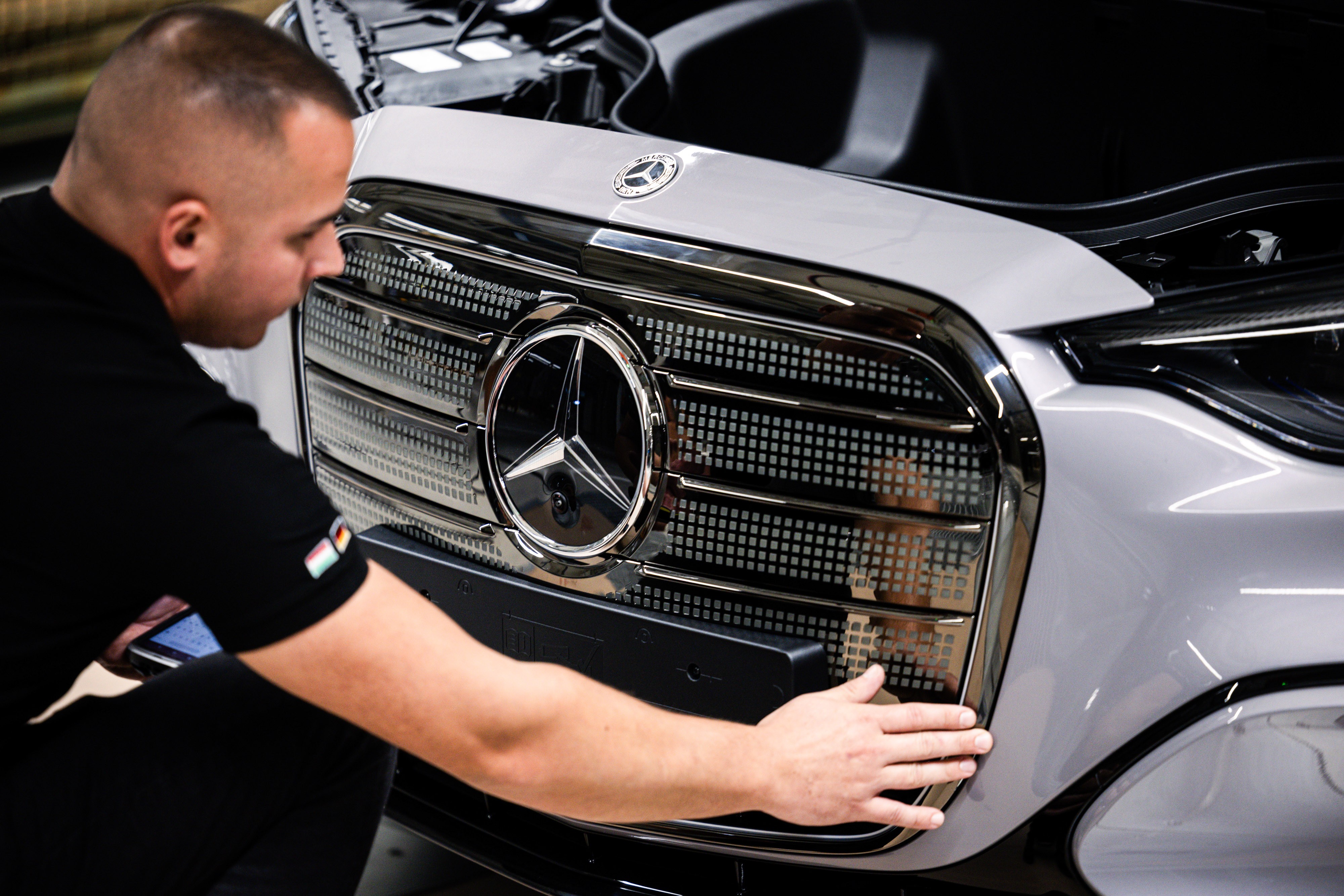 Homem de cabelo raspado, vestindo camiseta preta, inspeciona a grade frontal cromada de um carro Mercedes-Benz prateado, com o capô aberto