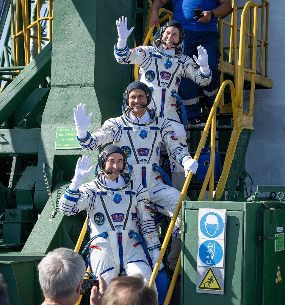 Três astronautas sorridentes, vestindo trajes espaciais brancos e azuis, acenam para a multidão enquanto descem uma escada metálica verde e amarela. Eles usam capacetes pretos com microfones e luvas brancas. Ao fundo, uma estrutura de lançamento de foguetes.