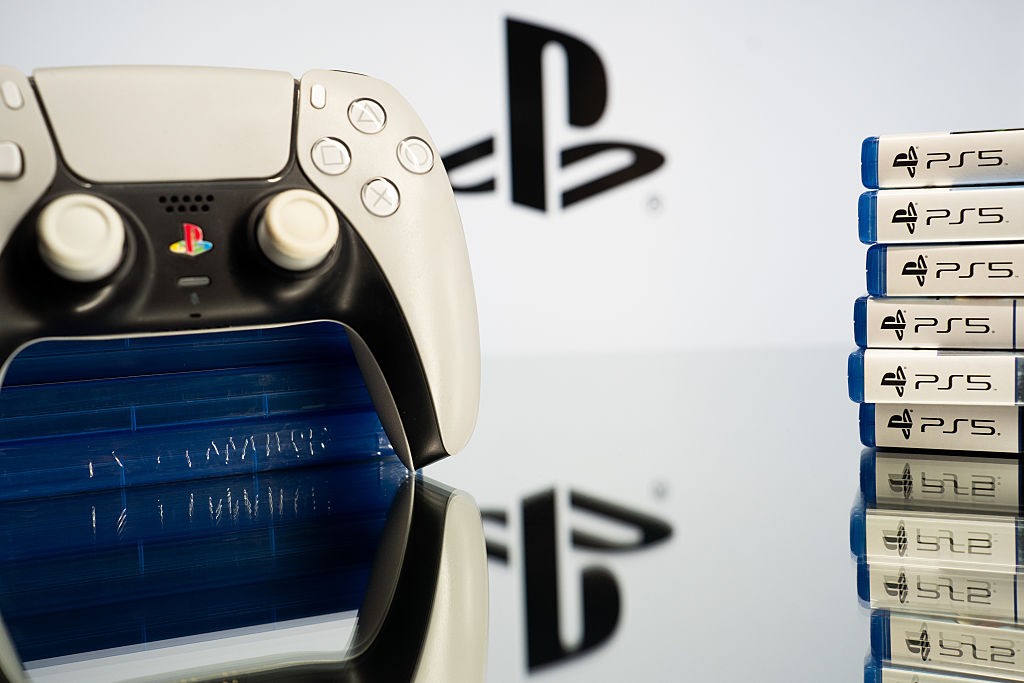 Controle branco e preto do PlayStation 5 com o logo colorido da marca, refletido em uma superfície escura. À direita, uma pilha de caixas de jogos PS5 com bordas azuis e o logo em preto, sobre um fundo branco com o logo PlayStation desfocado