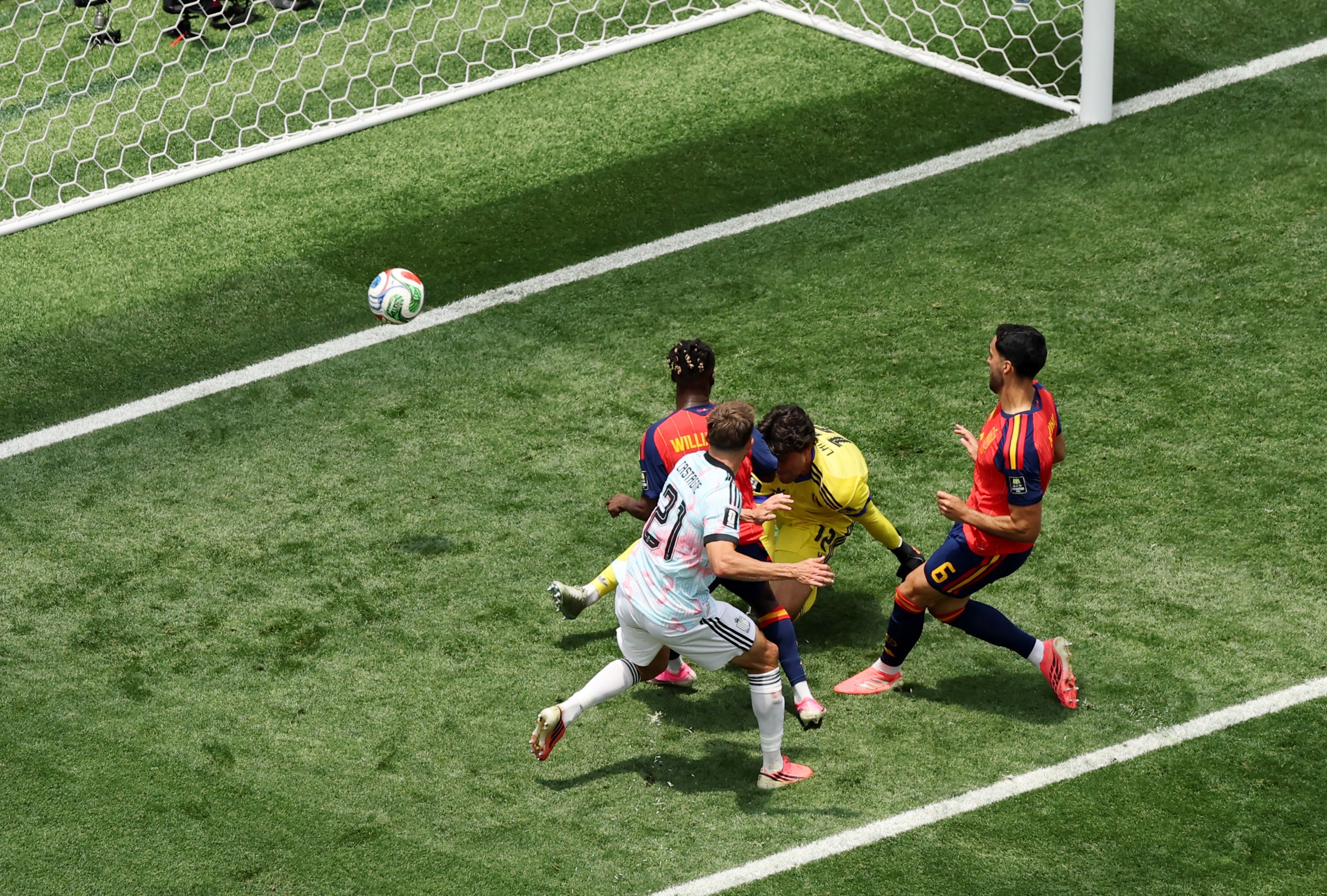 Quatro jogadores de futebol em campo verde, próximos ao gol. Um goleiro de camisa amarela se agacha, enquanto um jogador de camisa branca e preta chuta a bola colorida. Outros dois jogadores, um de camisa vermelha e azul e outro de camisa laranja e azul, observam a jogada. A rede do gol é visível no canto superior esquerdo