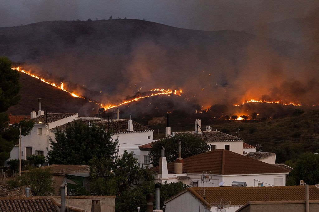 Incêndio florestal noturno em uma montanha, com chamas alaranjadas e fumaça escura, acima de casas brancas com telhados de telha marrom em primeiro plano