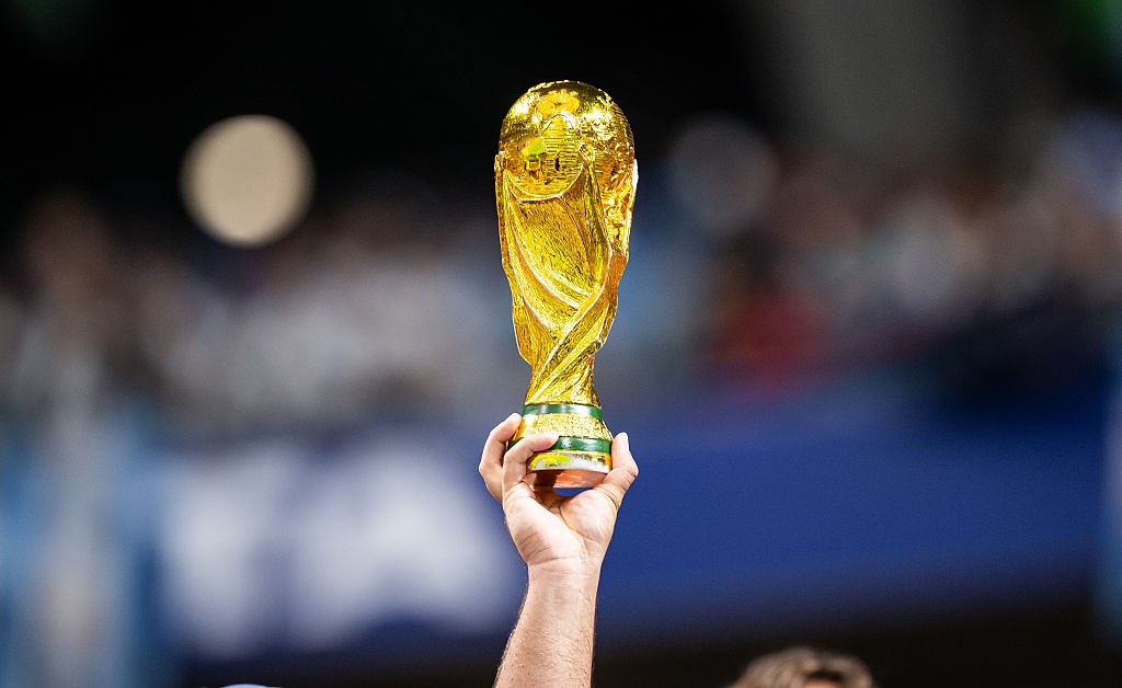 Mão de um homem erguendo a taça dourada da Copa do Mundo, com base verde, em um fundo desfocado de estádio e luzes