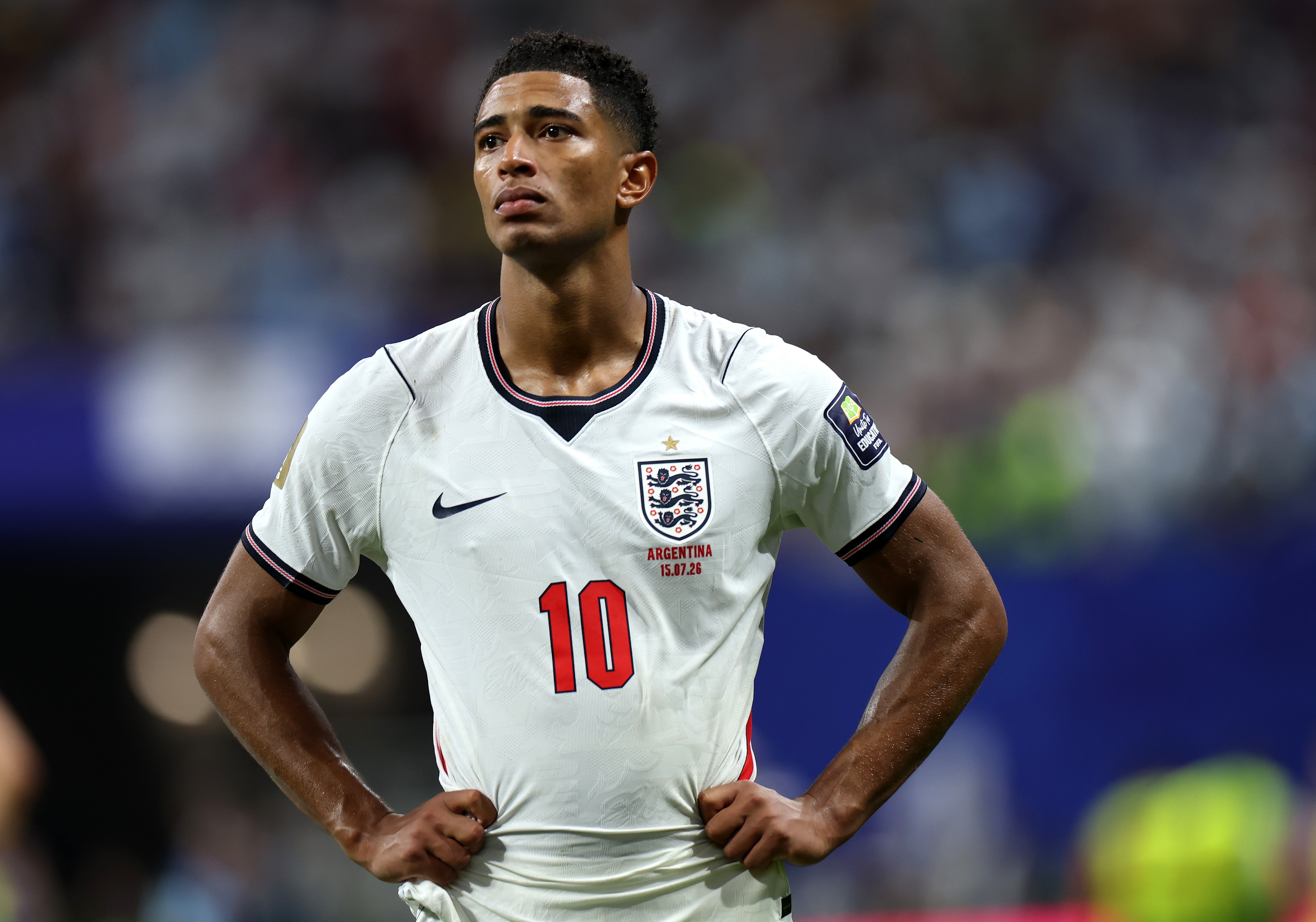 Jude Bellingham, jogador de pele morena e cabelo curto, veste camisa branca da seleção inglesa com o número 10 vermelho e o escudo do time. Ele está em campo, com as mãos na cintura e expressão séria, olhando para cima e para a direita. O fundo está desfocado, com tons de azul e branco, sugerindo um estádio lotado