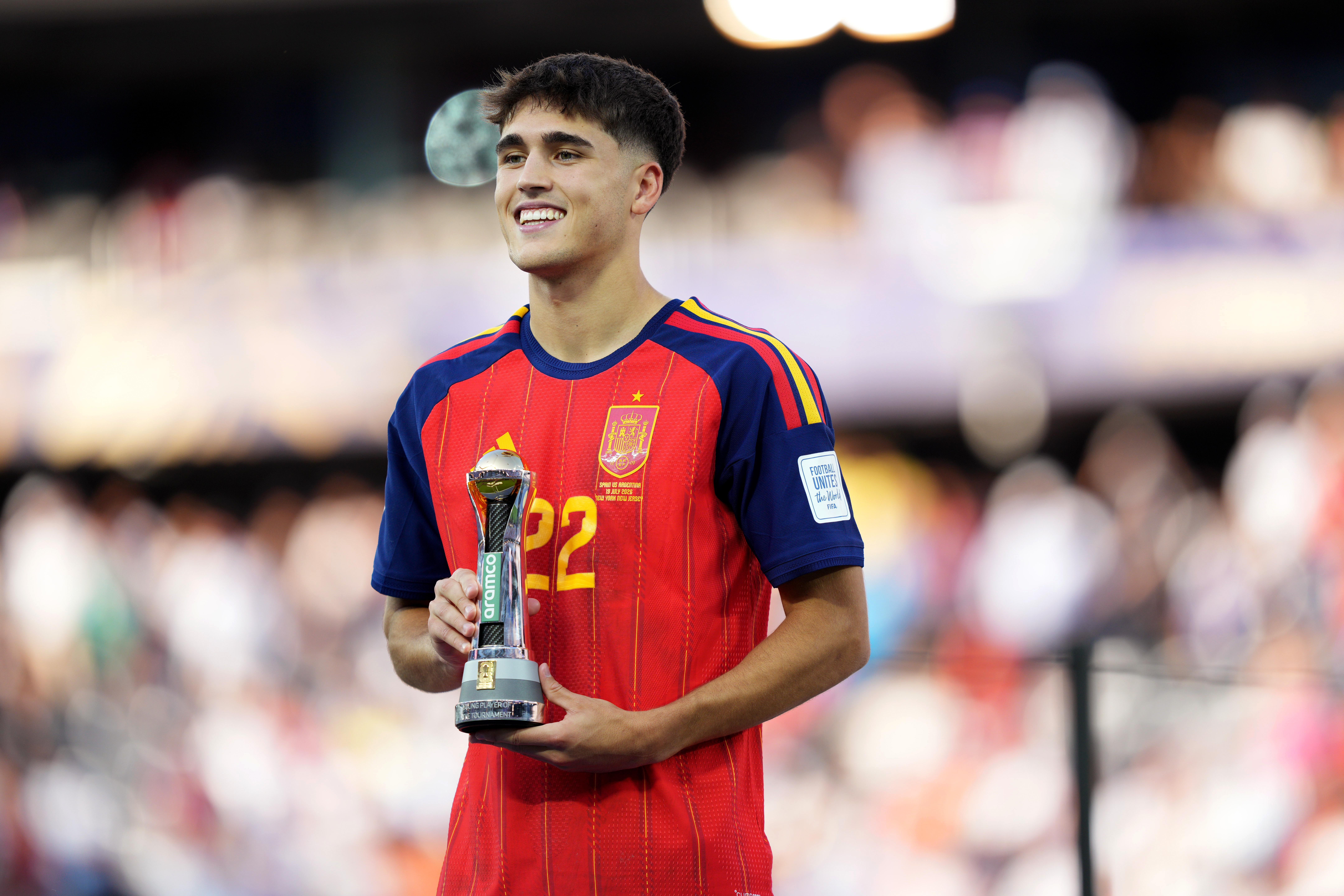Jovem jogador de futebol, com cabelo escuro e sorriso largo, vestindo camisa vermelha e azul da Espanha, segurando um troféu prateado com detalhes verdes, em um estádio com público desfocado ao fundo