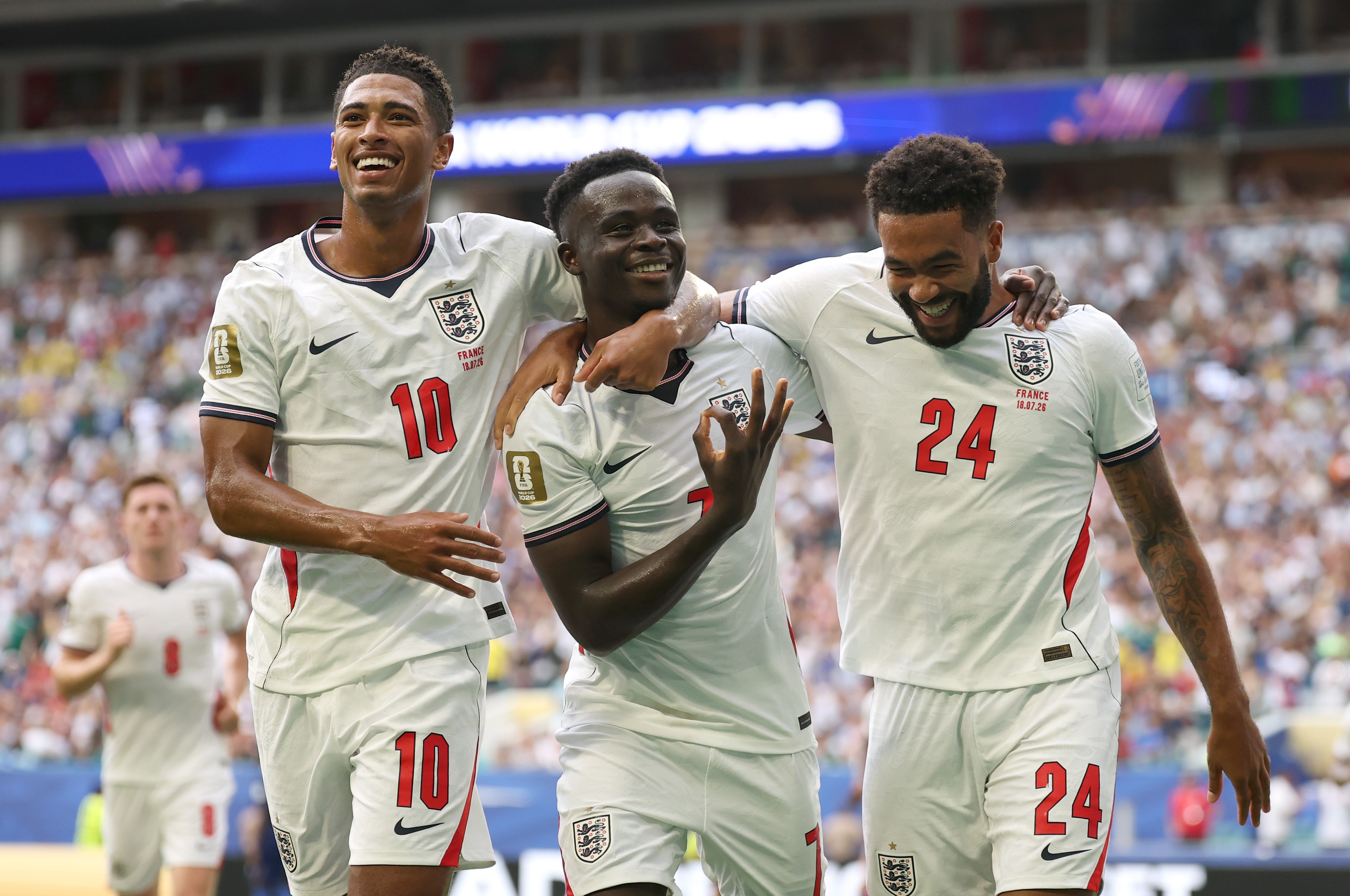 Três jogadores de futebol com camisas brancas da Inglaterra, sorrindo e se abraçando em campo. O jogador à esquerda, com a camisa 10, olha para cima. O do centro, com a camisa 7, faz um gesto de ok com a mão direita. O da direita, com a camisa 24, ri com a cabeça baixa.