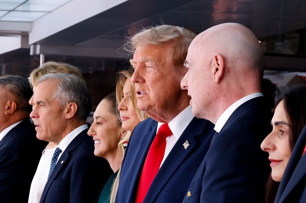 Várias pessoas em perfil, olhando para a direita. No centro, Donald Trump com terno azul e gravata vermelha, ao lado de um homem careca de terno azul. À esquerda, um homem de cabelos grisalhos e uma mulher sorrindo. À direita, uma mulher de cabelos escuros.