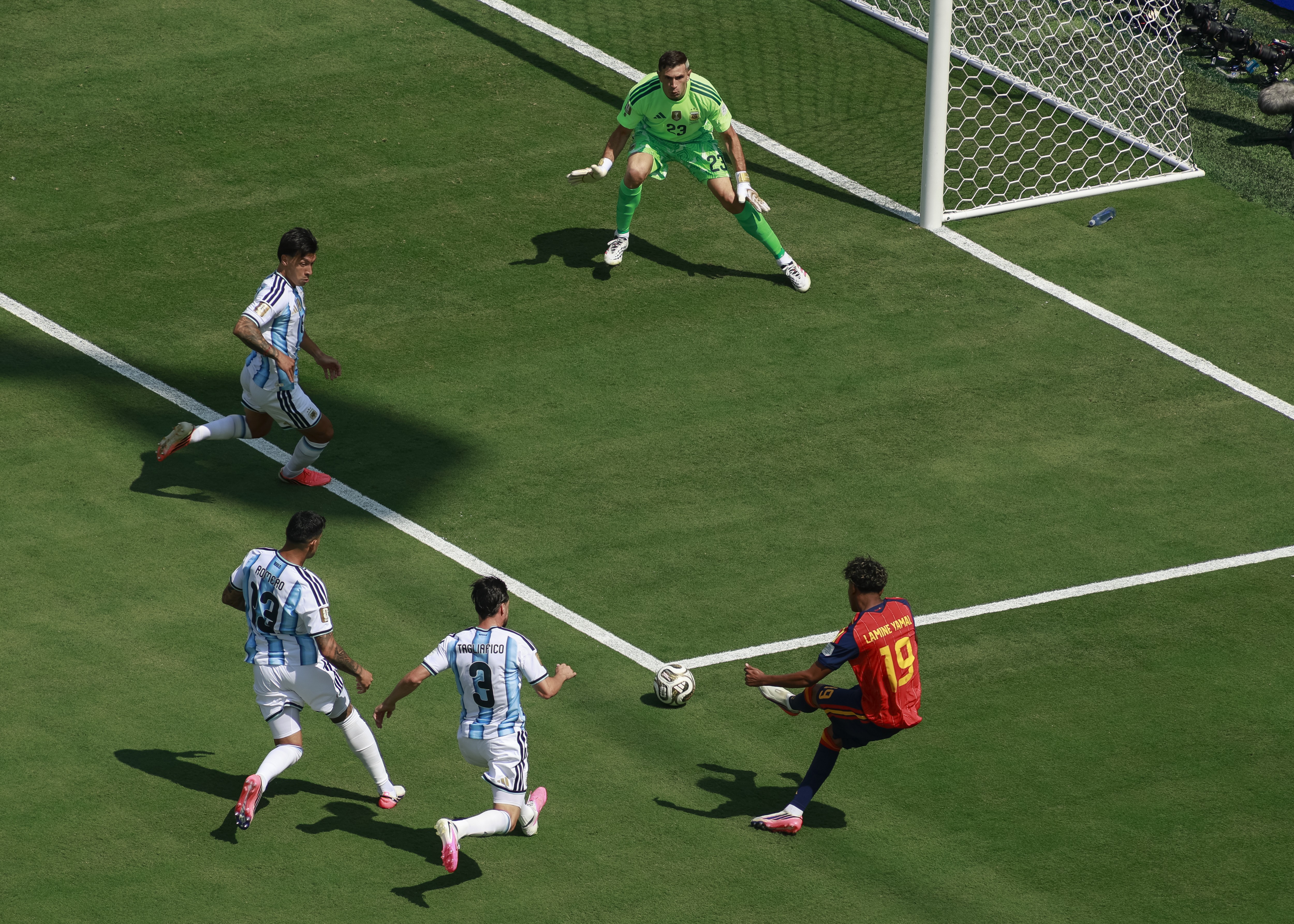 Um jogador de camisa vermelha com o número 19 chuta uma bola de futebol no gramado verde, enquanto três jogadores de camisa branca e azul listrada e um goleiro de camisa verde observam a jogada perto do gol