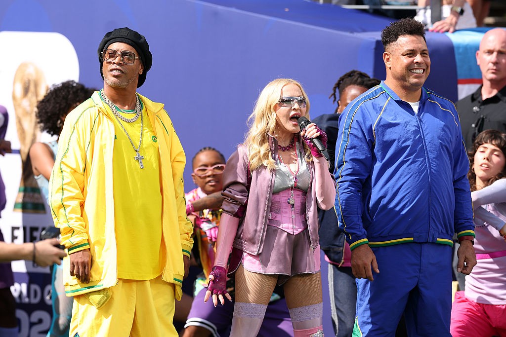 Ronaldinho Gaúcho de amarelo, Madonna de rosa cantando no microfone e Ronaldo Fenômeno de azul, sorrindo, em um palco com fundo azul
