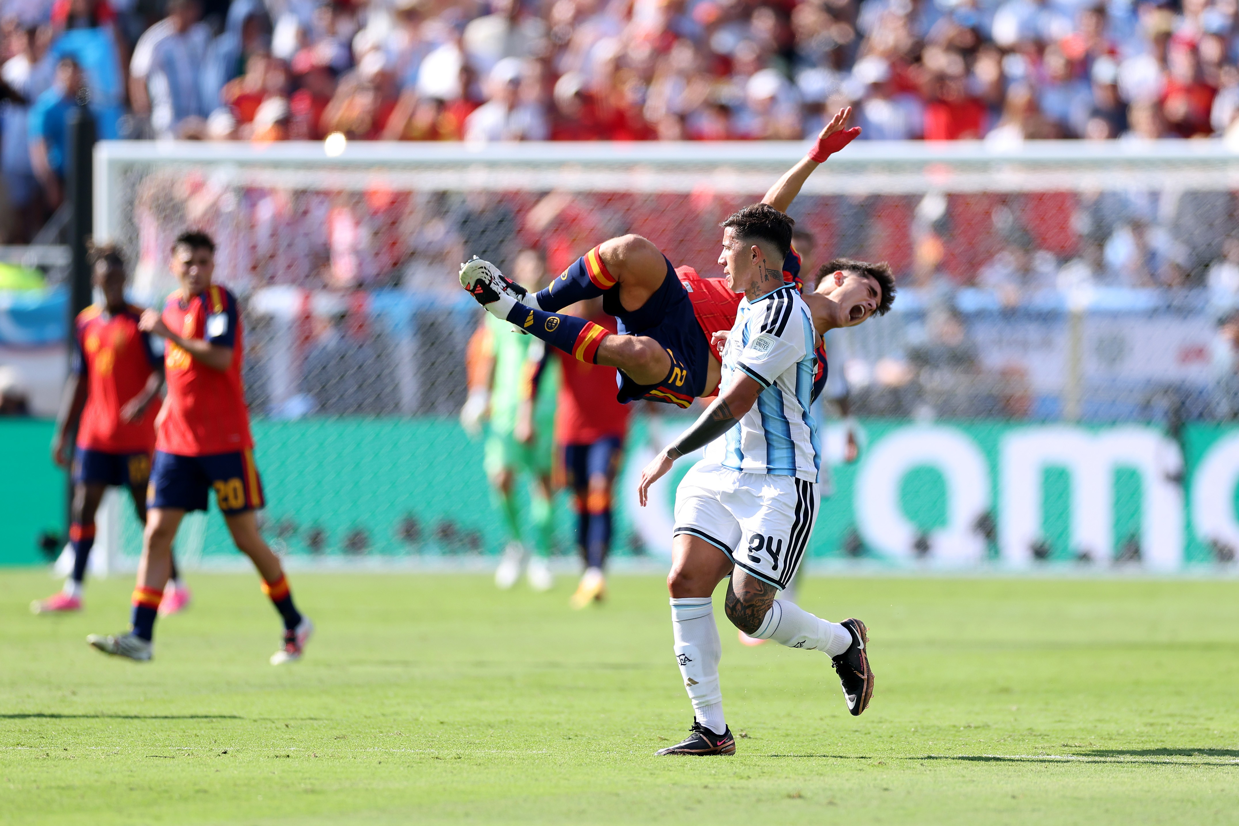Dois jogadores de futebol em campo. Um, de camisa azul e branca, está de pé, olhando para o outro, que está no ar, de cabeça para baixo, com a camisa vermelha e shorts azuis, chutando a bola. Outros jogadores e torcedores desfocados ao fundo