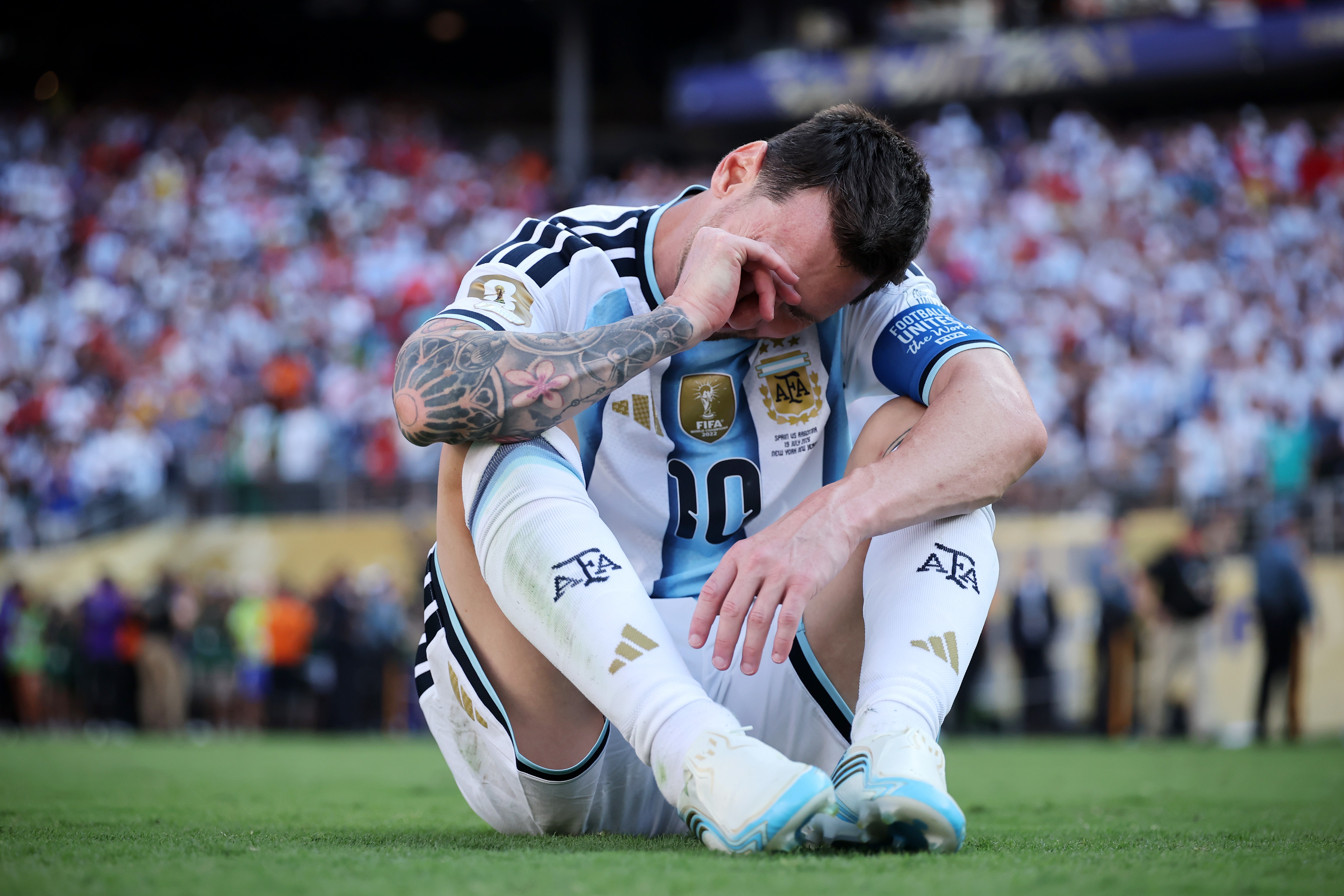 Jogador de futebol Lionel Messi, com camisa da Argentina, sentado no gramado com a cabeça baixa e mão no rosto, expressando tristeza. Ao fundo, torcida embaçada