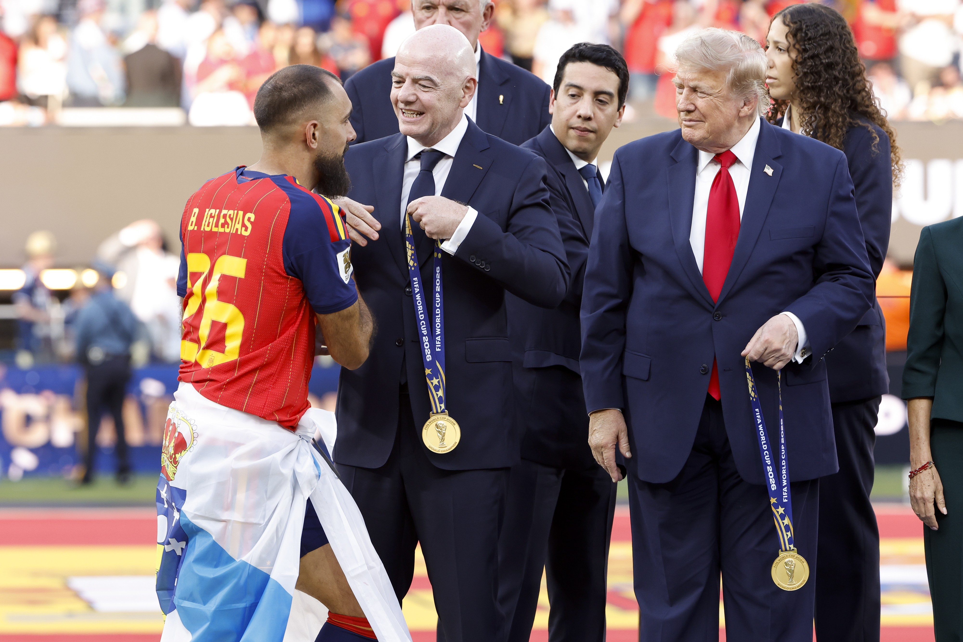 Um jogador de futebol com camisa vermelha e azul, número 26 e nome B. Iglesias, conversa com Gianni Infantino, presidente da FIFA, que sorri e segura uma medalha dourada. Ao lado, Donald Trump, de terno azul e gravata vermelha, também segura uma medalha, olhando para a esquerda.