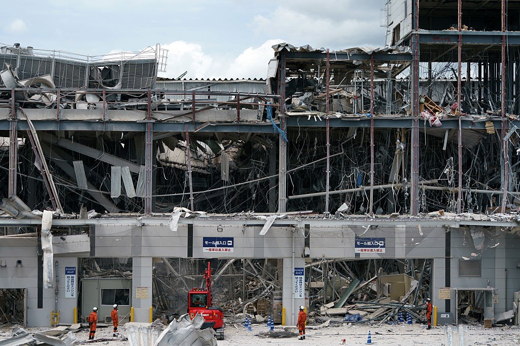 Edifício de vários andares com estrutura metálica exposta e escombros, indicando destruição. Trabalhadores de laranja e uma máquina vermelha estão no térreo, em meio aos destroços