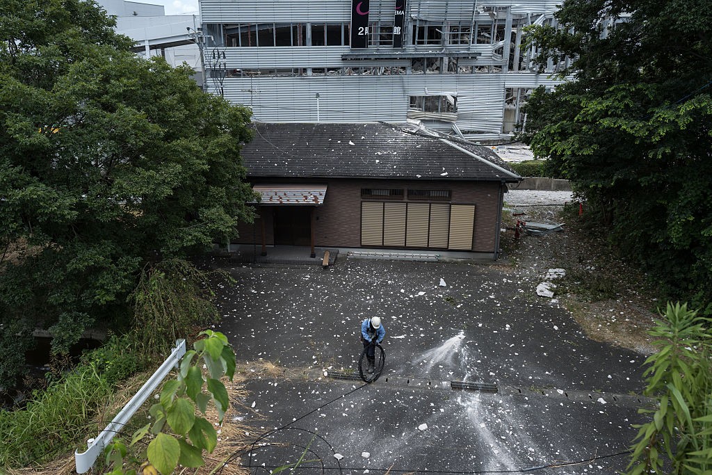 Vista aérea de um homem de capacete e roupa azul limpando um pátio escuro com uma mangueira de alta pressão, espalhando detritos brancos. Ao fundo, uma casa de telhado escuro e paredes de tijolos, e um prédio maior, parcialmente destruído, com janelas quebradas e estrutura metálica exposta. Árvores verdes densas cercam a cena, com vegetação rasteira à esquerda e direita