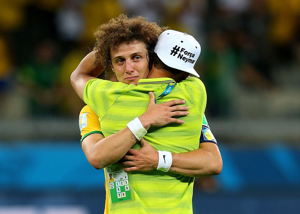 Dois jogadores de futebol brasileiros se abra&ccedil;am em campo. Um deles, com cabelos cacheados e express&atilde;o triste, chora enquanto abra&ccedil;a o outro, que veste um bon&eacute; branco com a frase #For&ccedil;aNeymar e tem as costas viradas para a c&acirc;mera. Ambos usam camisas amarelas e munhequeiras brancas, em um momento de consolo ap&oacute;s uma partida. O fundo est&aacute; desfocado, com tons escuros e alguns pontos de luz amarela