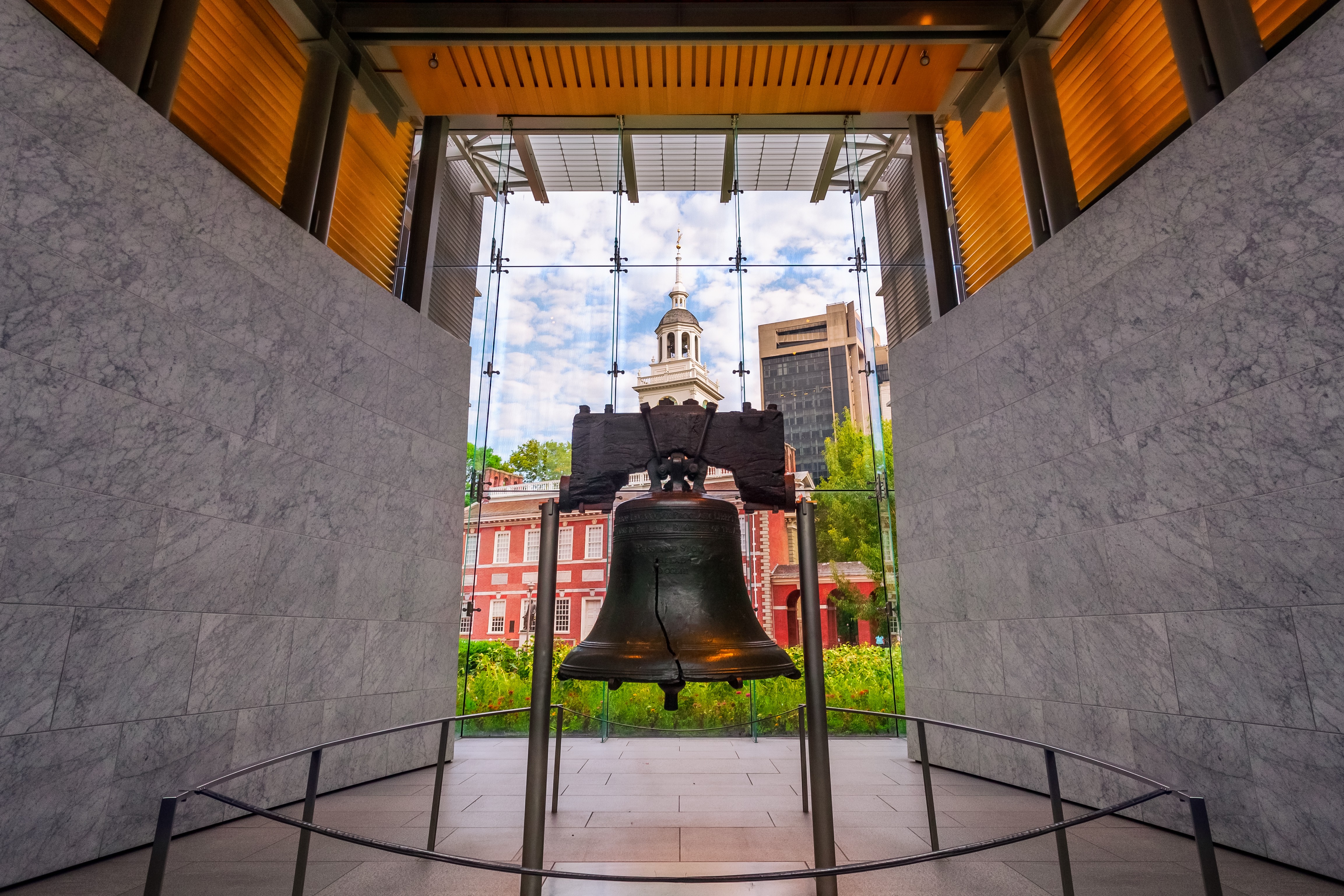 O Sino da Liberdade, rachado e escuro, em exposição dentro de uma estrutura moderna com paredes de mármore cinza e teto de madeira. Ao fundo, através de uma parede de vidro, vê-se o Independence Hall com sua torre branca e um céu azul com nuvens